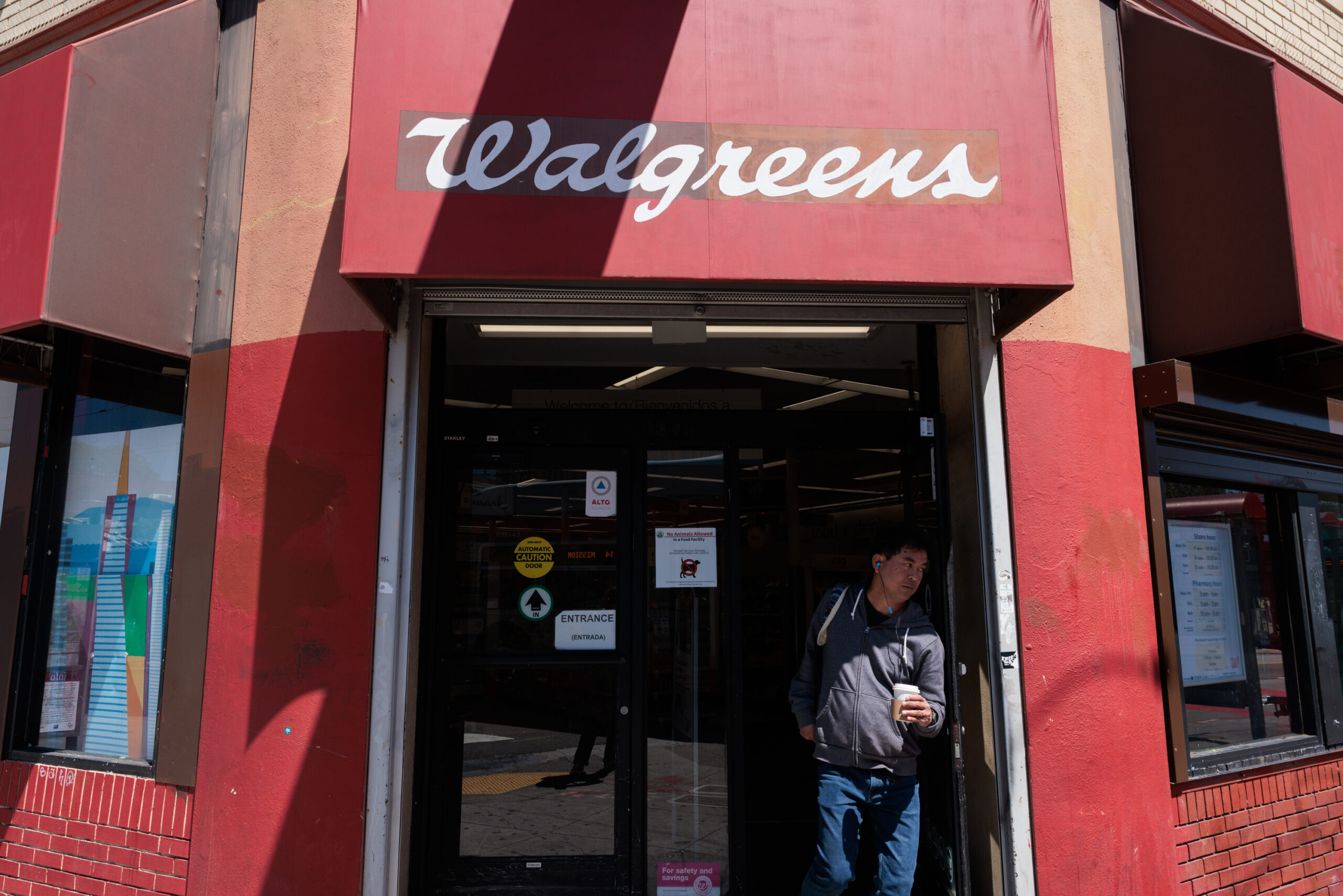 A person is exiting a Walgreens store, wearing a dark hoodie and carrying a drink. The storefront features a red awning with the Walgreens logo.