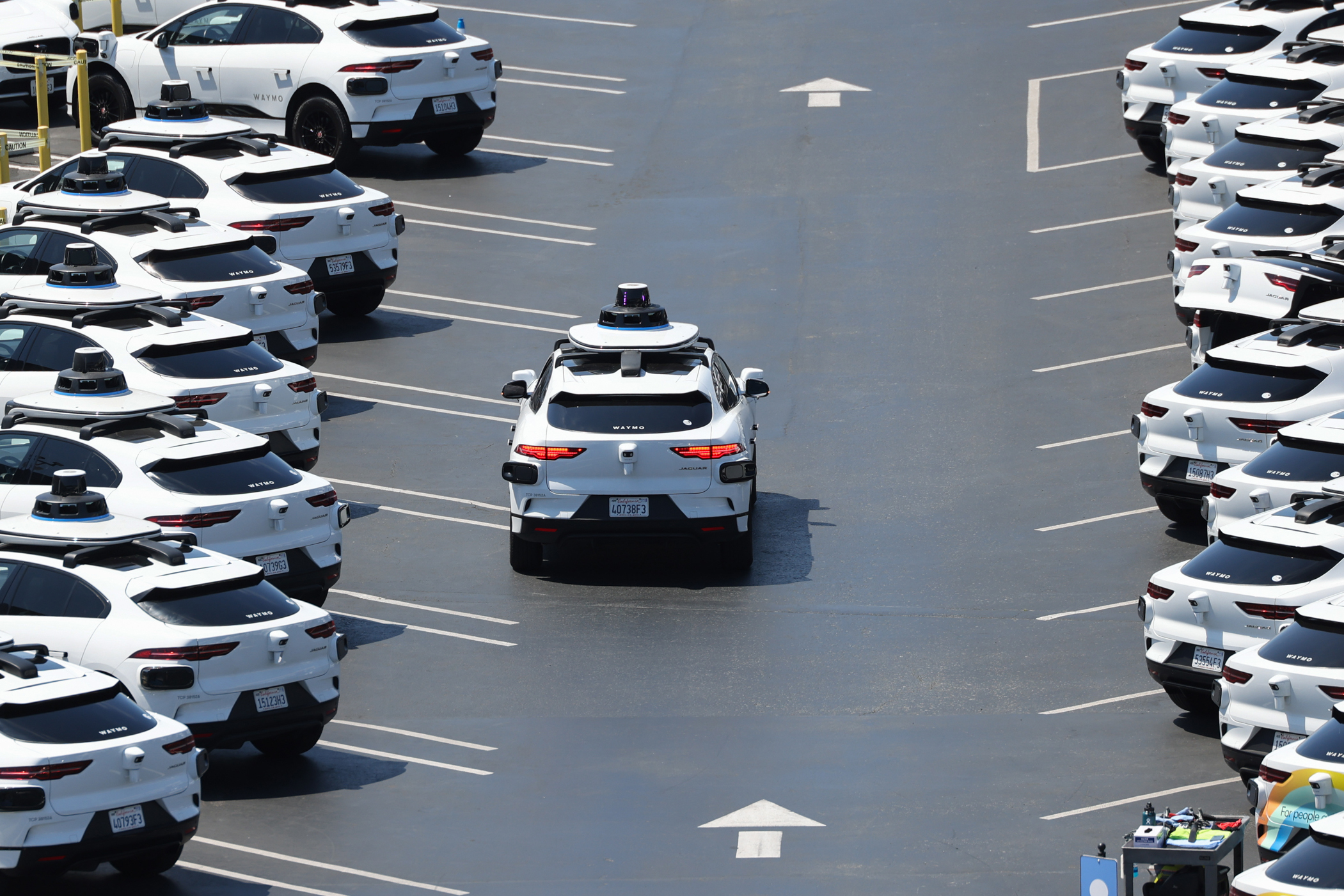 The image depicts numerous identical white SUVs equipped with sensors, parked in an organized manner in a parking lot, with one SUV driving down the central lane.