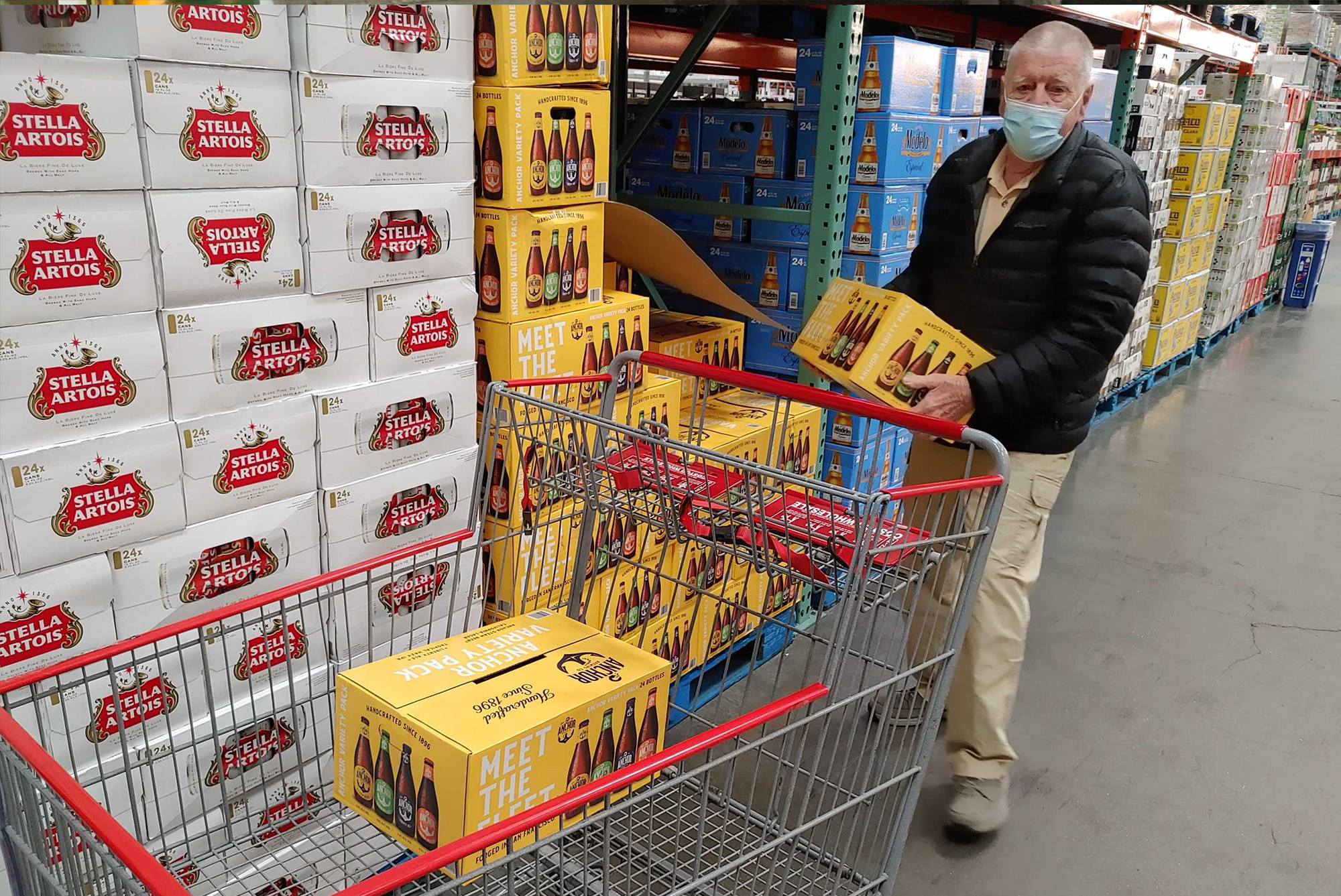 A person in a mask is placing a case of assorted beer bottles into a shopping cart in a store with stacks of Stella Artois and other beer brands.