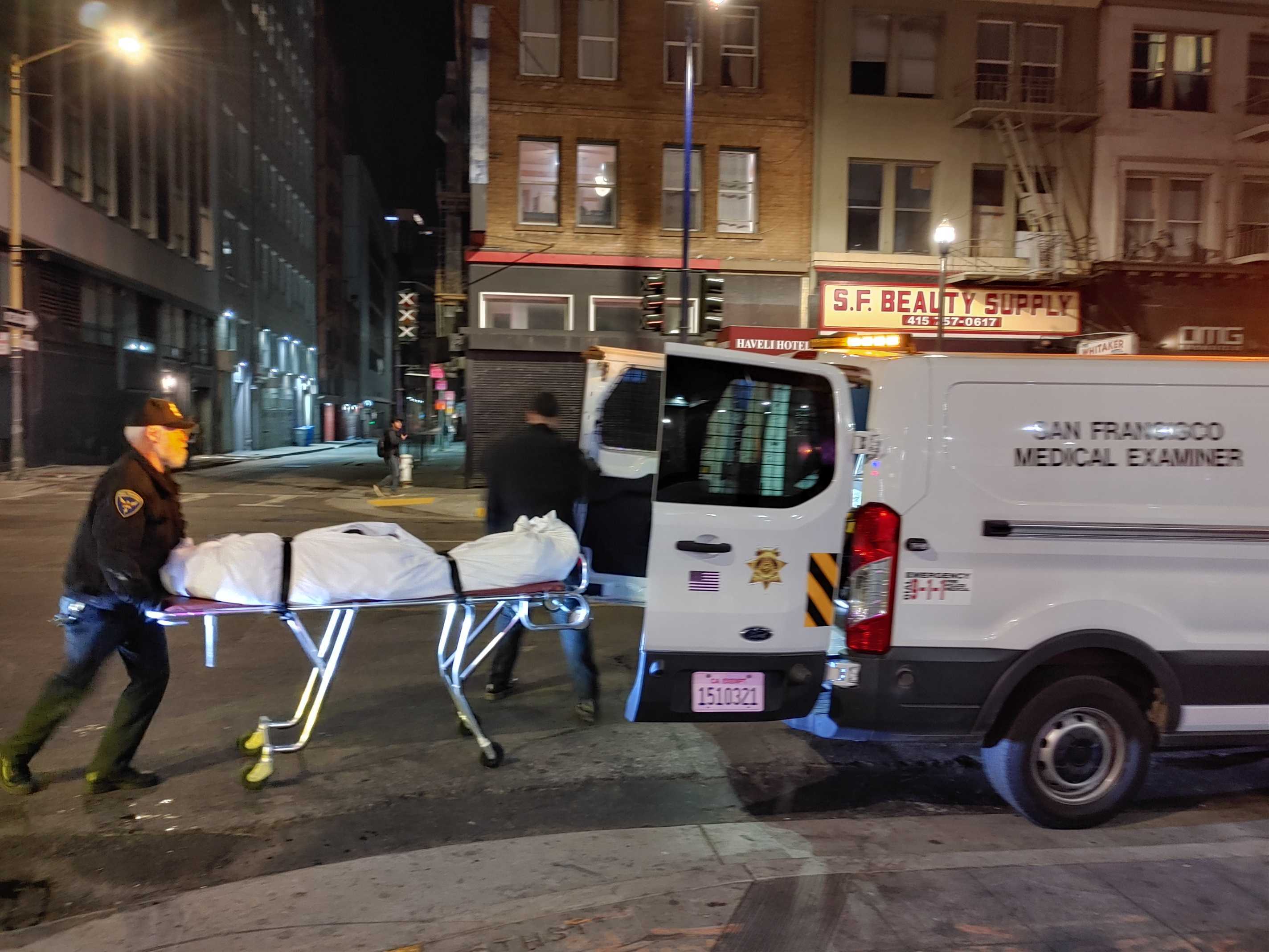 Officials are loading a covered stretcher into a medical examiner's van at night on a city street.