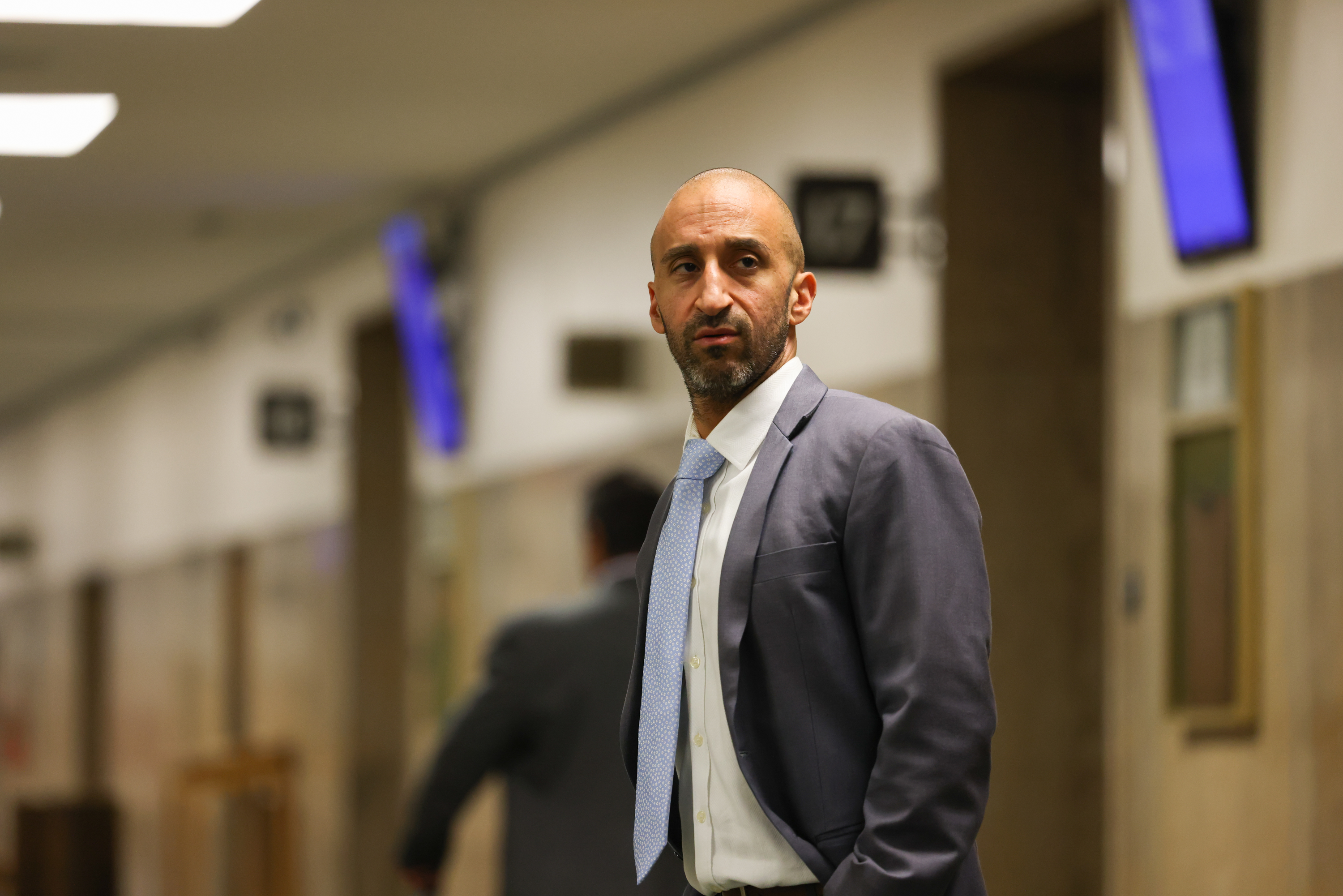 A man in a suit and tie stands in a hallway with beige walls and blurred background screens. He appears focused and slightly serious, looking off to the side.