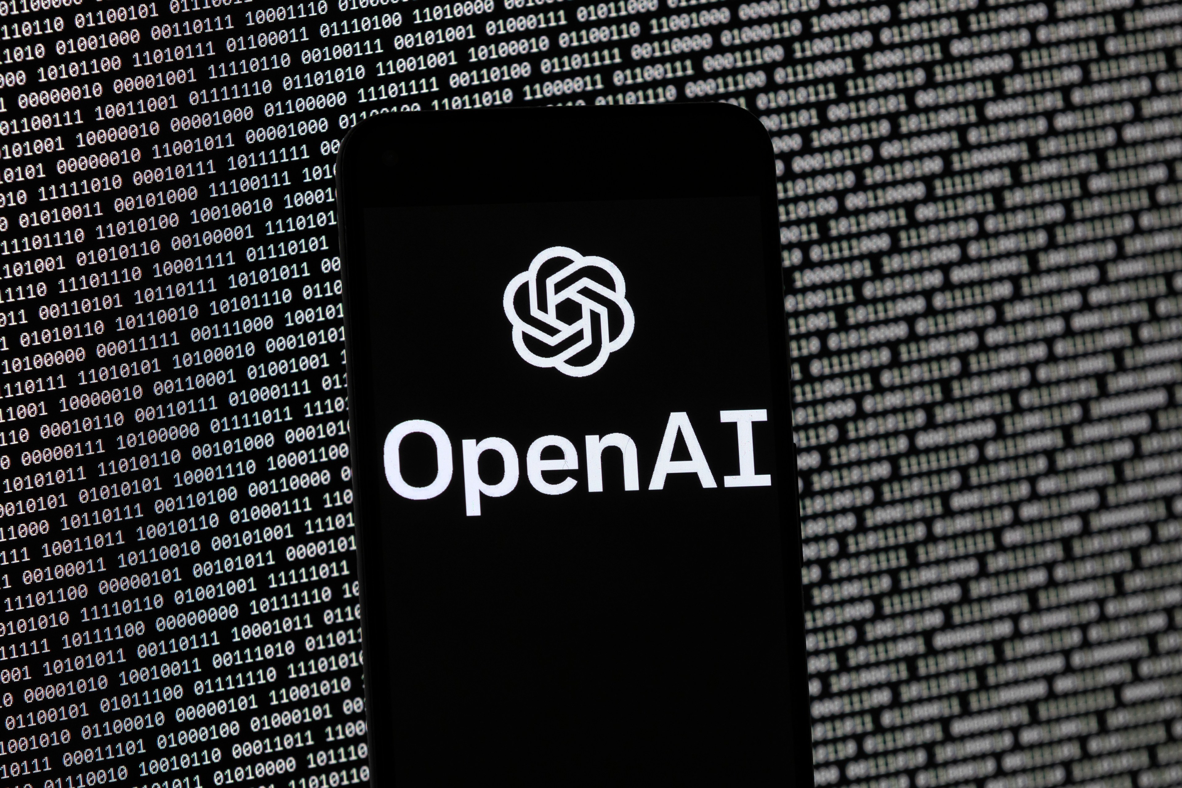 The logo of the Open Ai logo on a mobile device with aa series of zeros and ones in the background on a monitor