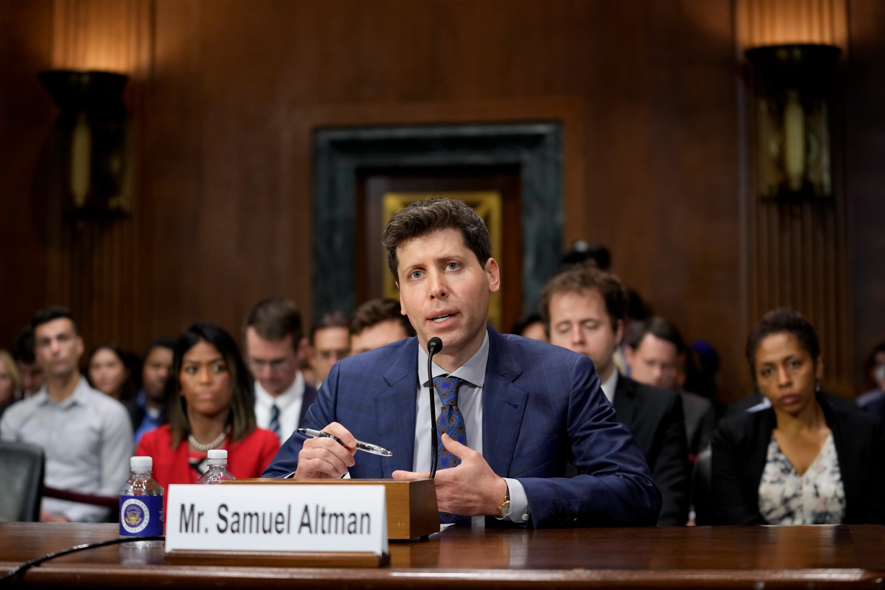 A man in a suit speaks at a hearing, people behind him, "Mr. Samuel Altman" sign in front.