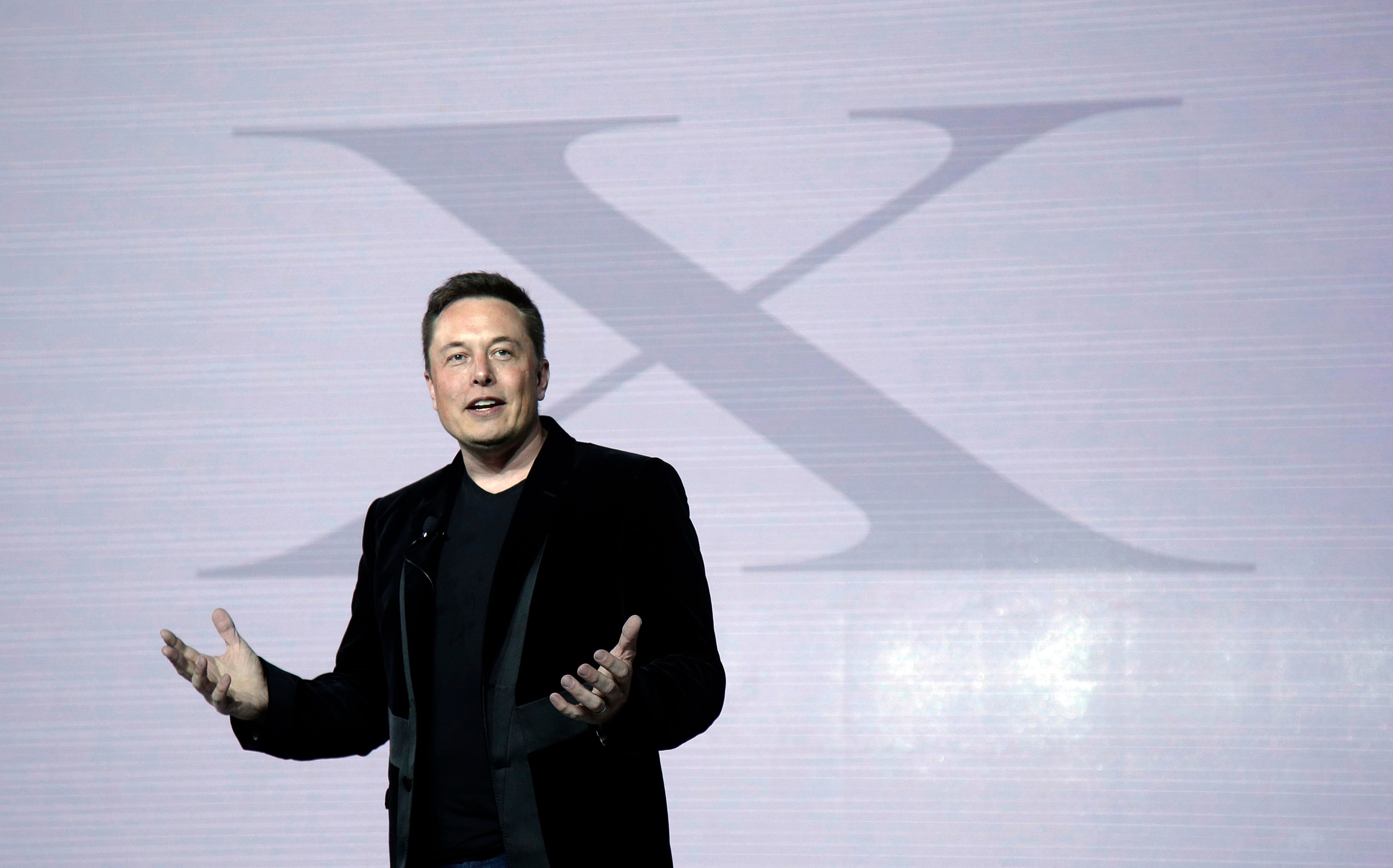 Elon Musk stands on a state with X in the background.