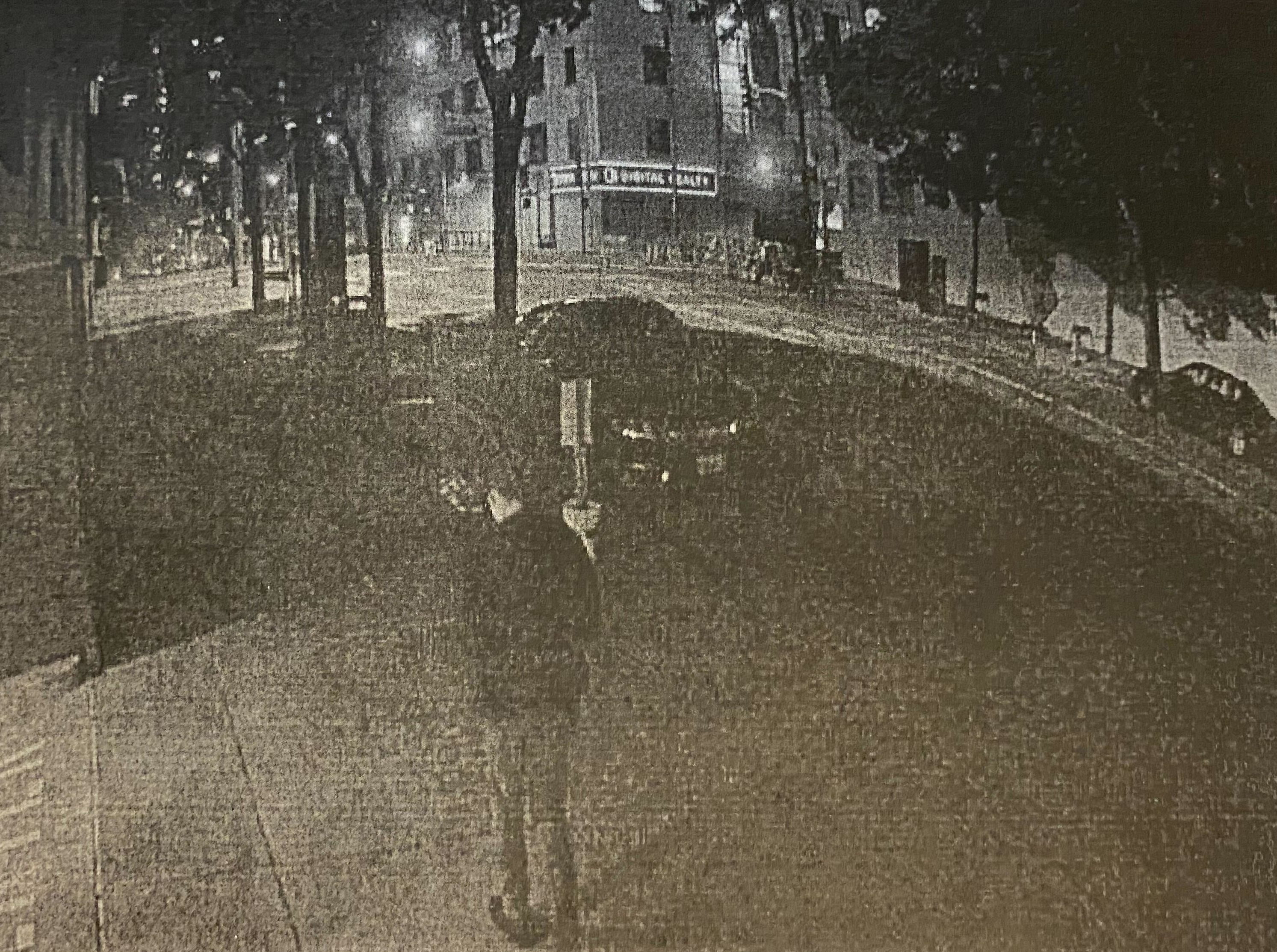 A grainy, black-and-white nighttime street scene shows a person walking on a sidewalk under streetlights near parked cars and trees.