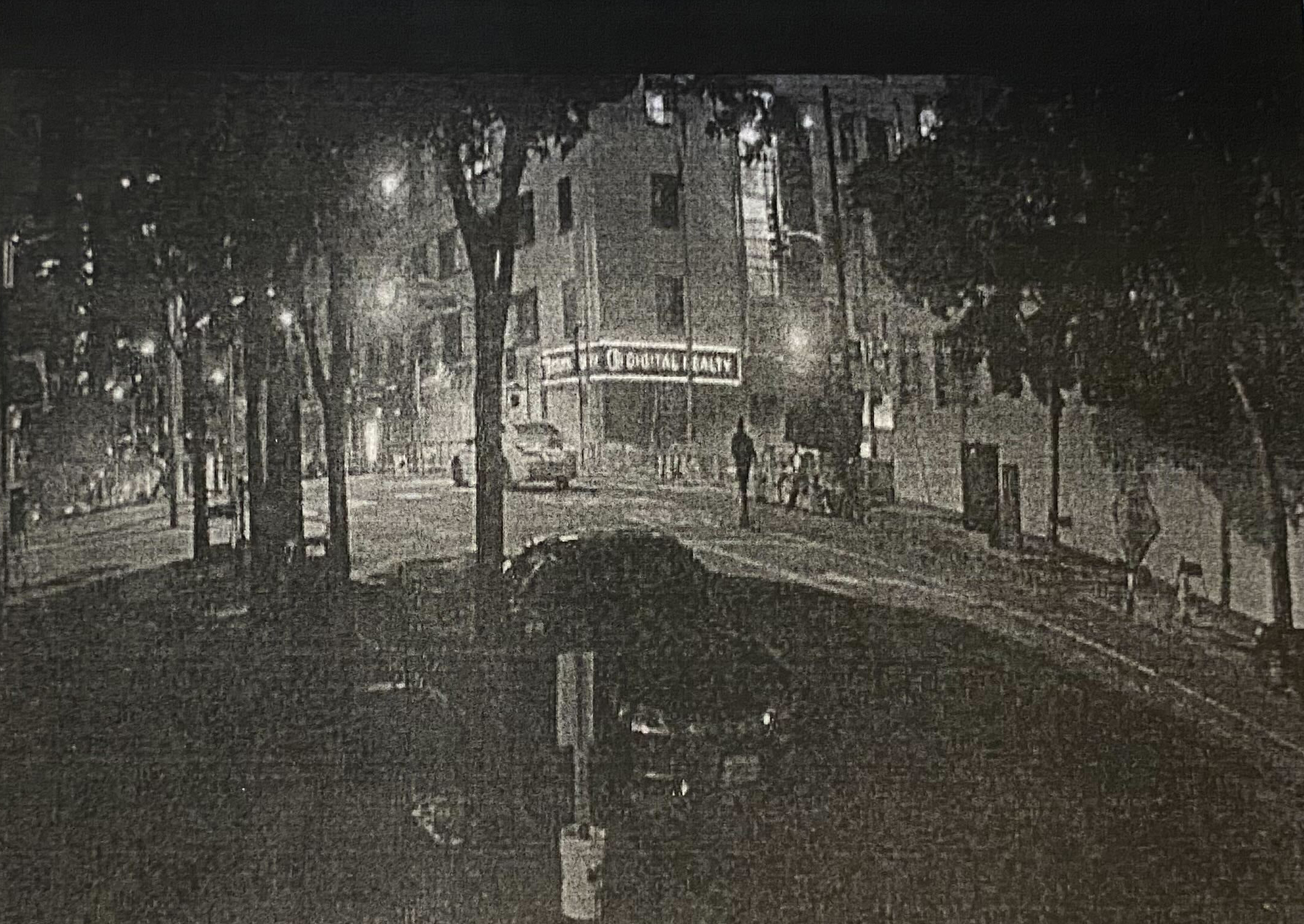 The black-and-white image shows a quiet, dimly-lit urban street at night with parked cars, trees lining the sidewalk, and a distant pedestrian. A building sign reads &quot;Digital Realty.&quot;