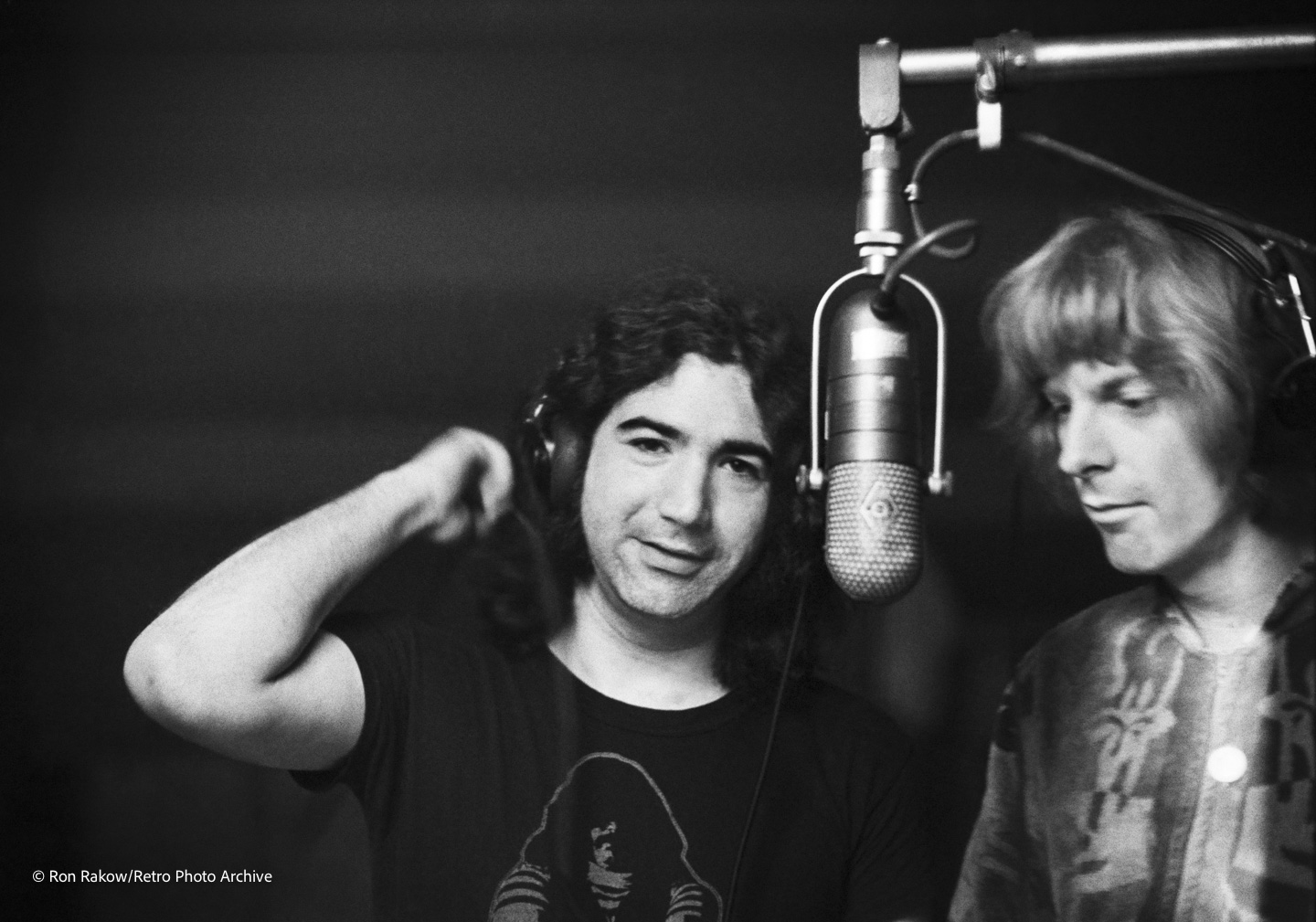 Two men are in a recording studio near a large microphone. One is looking at the camera while wearing headphones, and the other is looking down with an unreadable expression.