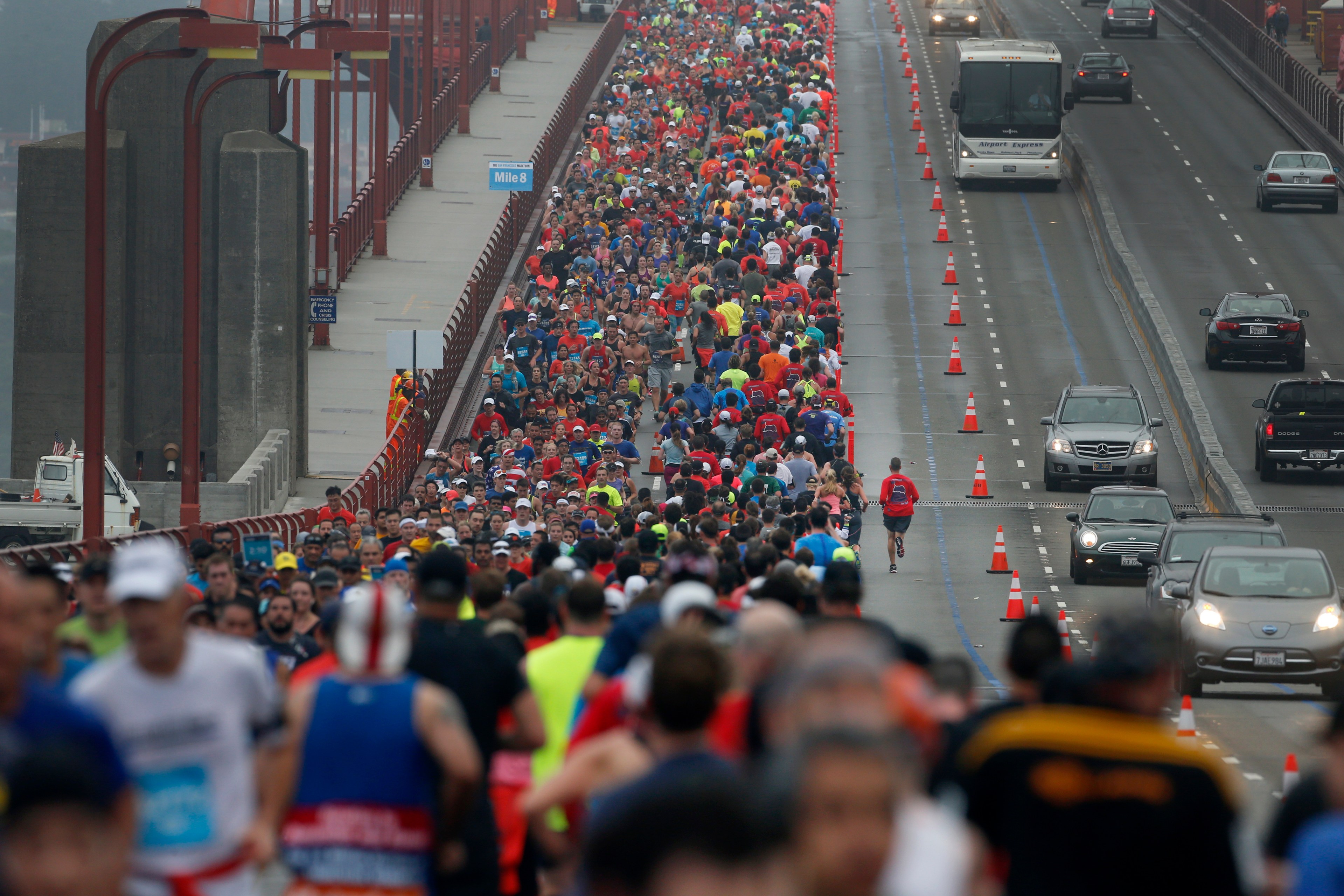 A large crowd of runners, some in brightly colored attire, participate in a race on a bridge. Traffic is separated from the runners by cones on the right.