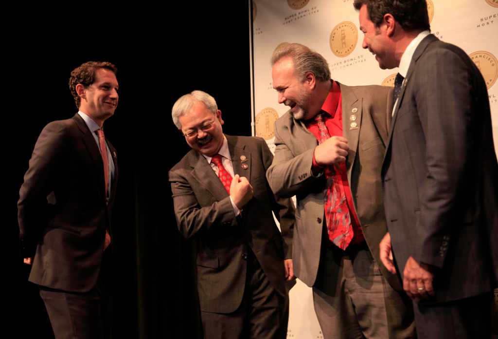 Daniel Lurie, San Francisco Mayor Ed Lee and two other men in suits share a lighthearted moment on stage for Super Bowl 50 preparations.