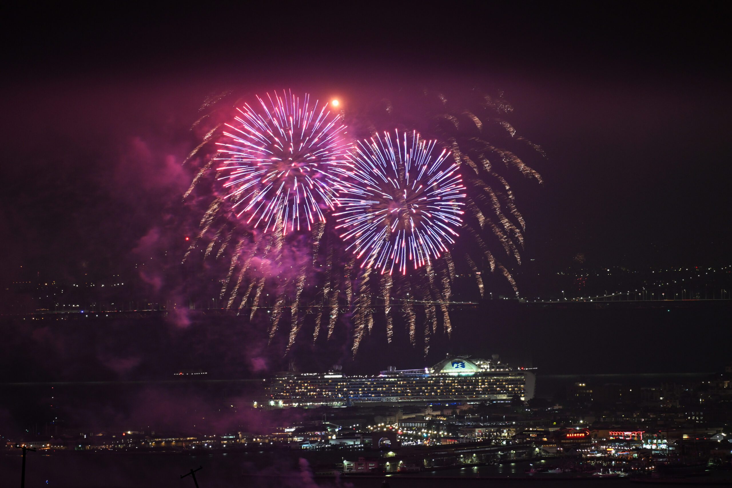 Bright fireworks burst in the night sky over a lit-up cruise ship and a cityscape, with a bridge in the background, creating a festive atmosphere.