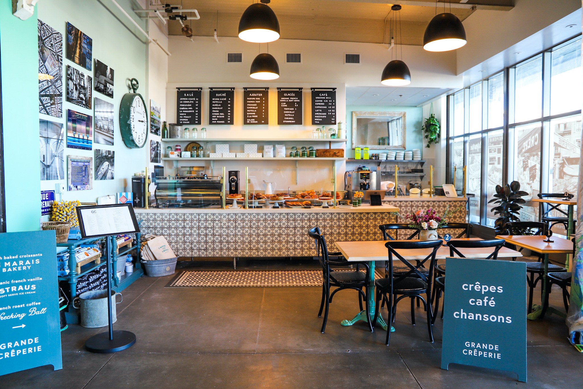 The image shows a bright café with patterned counter tiles, wall art, hanging lights, and large windows. Tables with chairs are in the foreground.