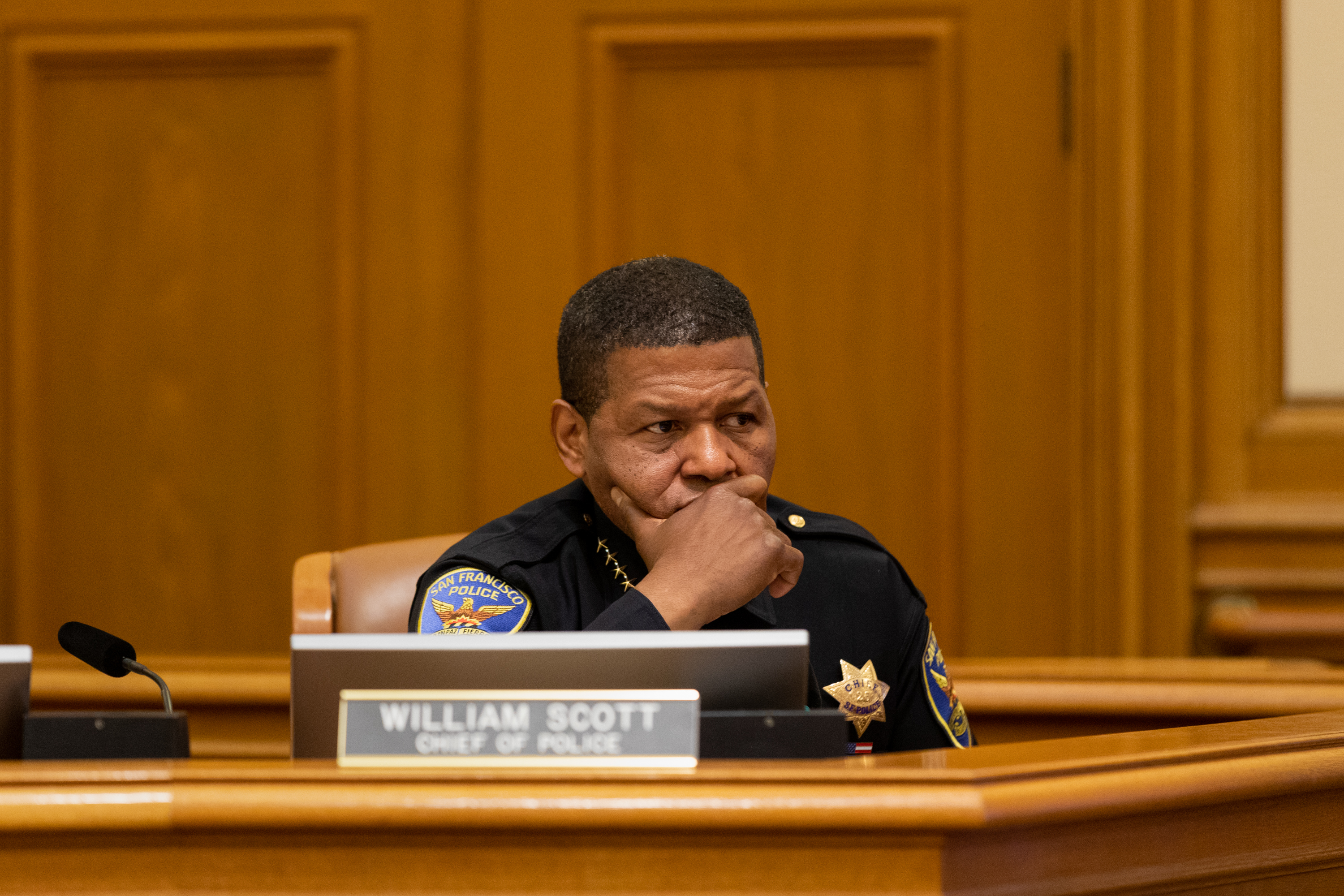 A man in a police uniform sits thoughtfully at a desk with a microphone and a nameplate reading &quot;WILLIAM SCOTT, CHIEF OF POLICE.&quot;