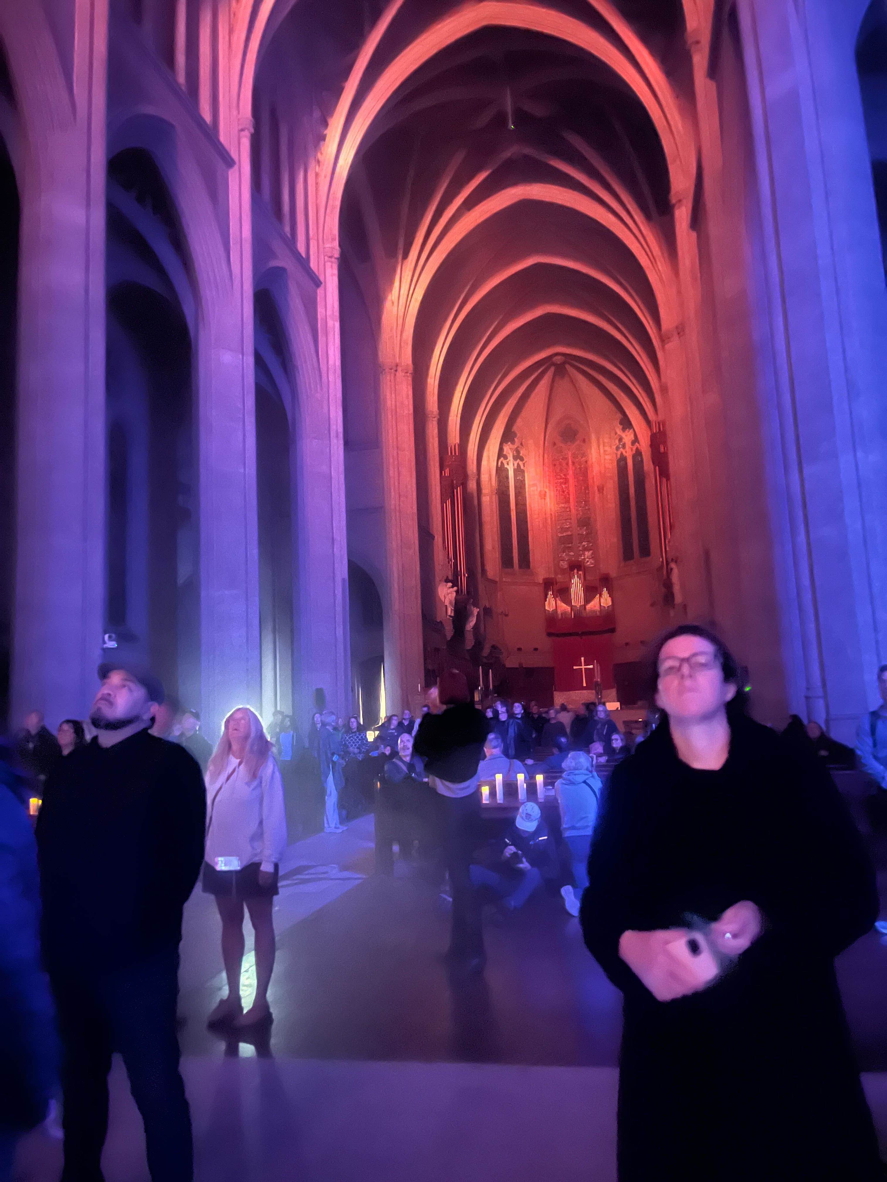 People are standing in a large, dimly lit cathedral with tall arches and colorful lighting. An altar and large cross are visible in the background.