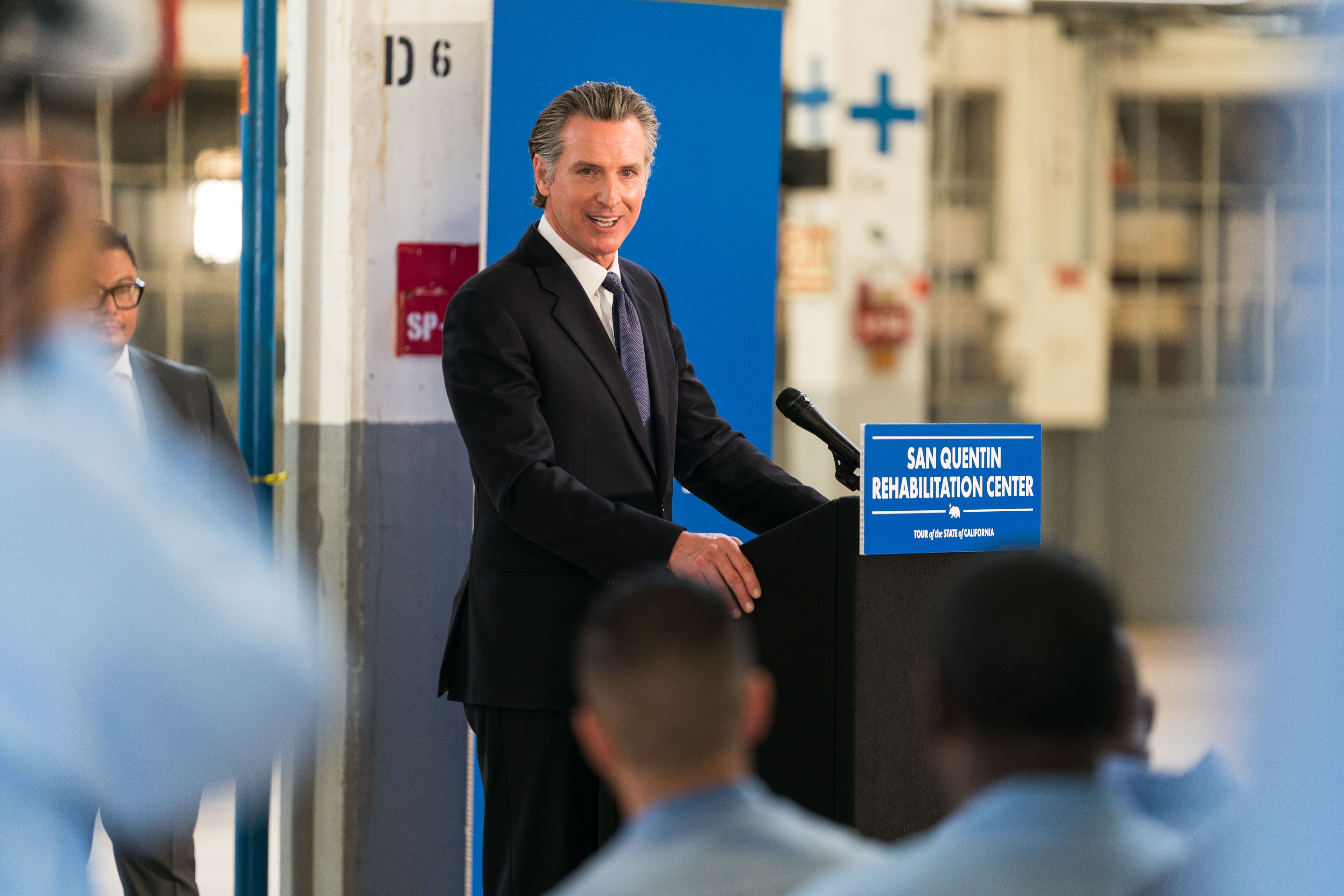 A man in a suit stands at a podium marked &quot;San Quentin Rehabilitation Center&quot; with blurred listeners in the foreground.