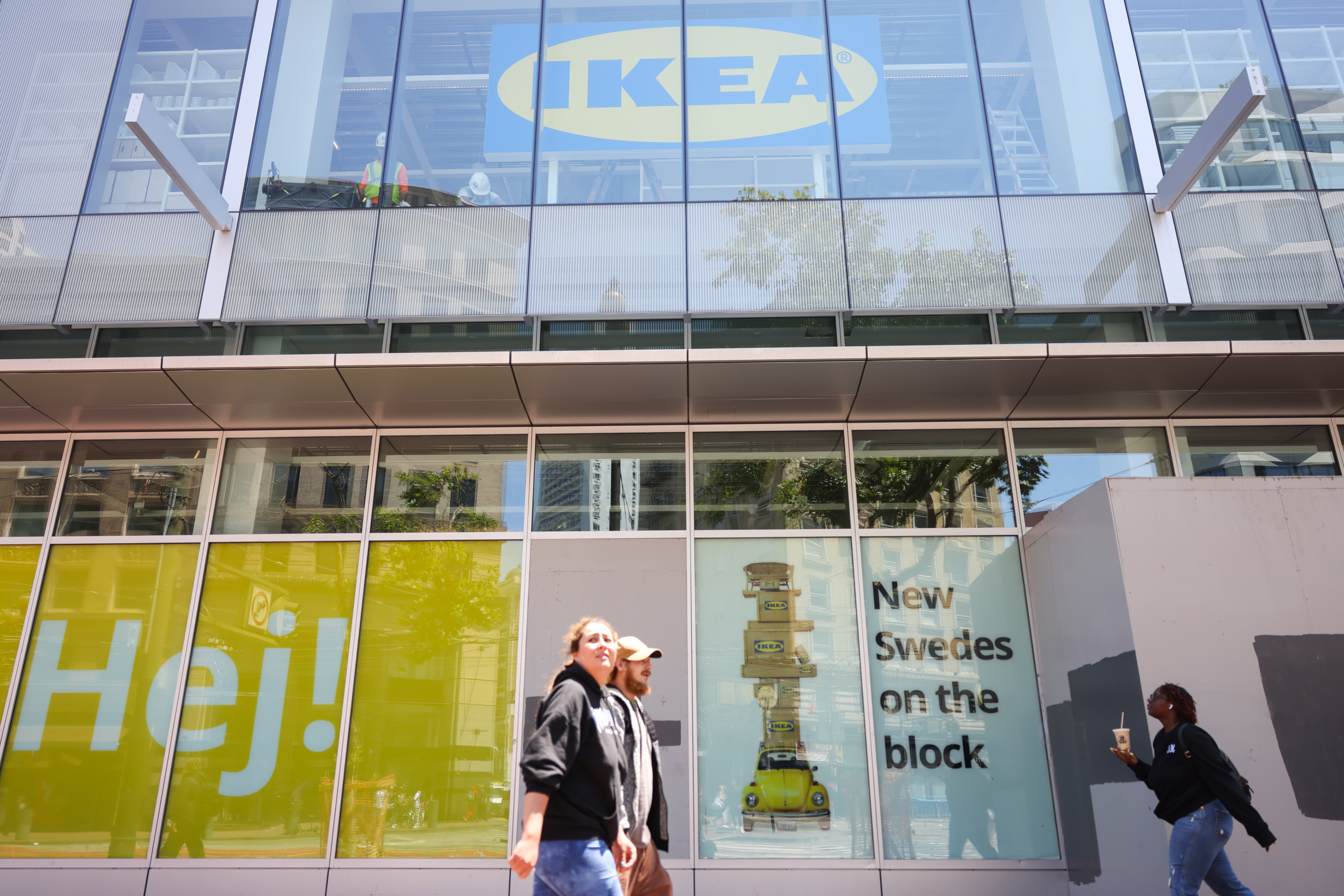 Two people walk by a glass-front IKEA store displaying the logo and quirky text "New Swedes on the block."
