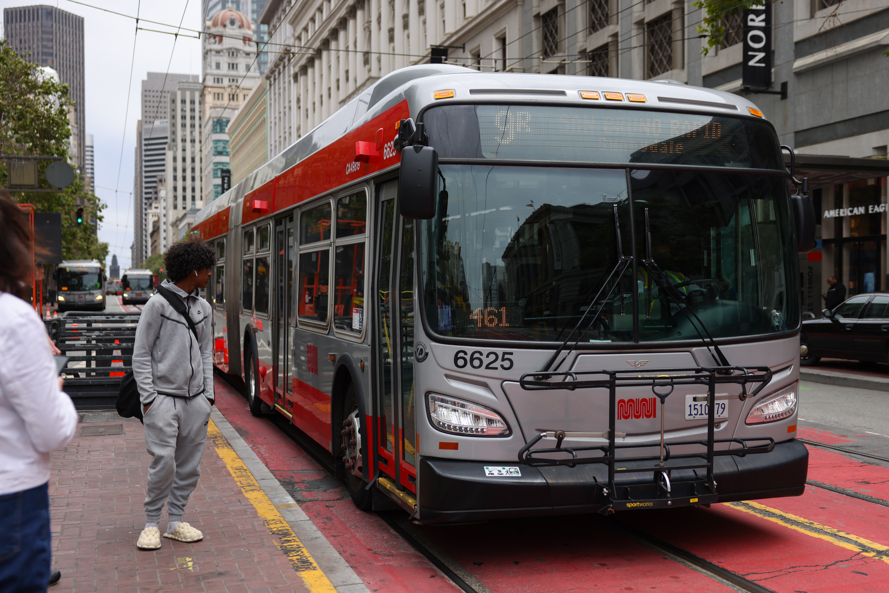 A red city bus with a digital sign is parked on a street, with pedestrians nearby.