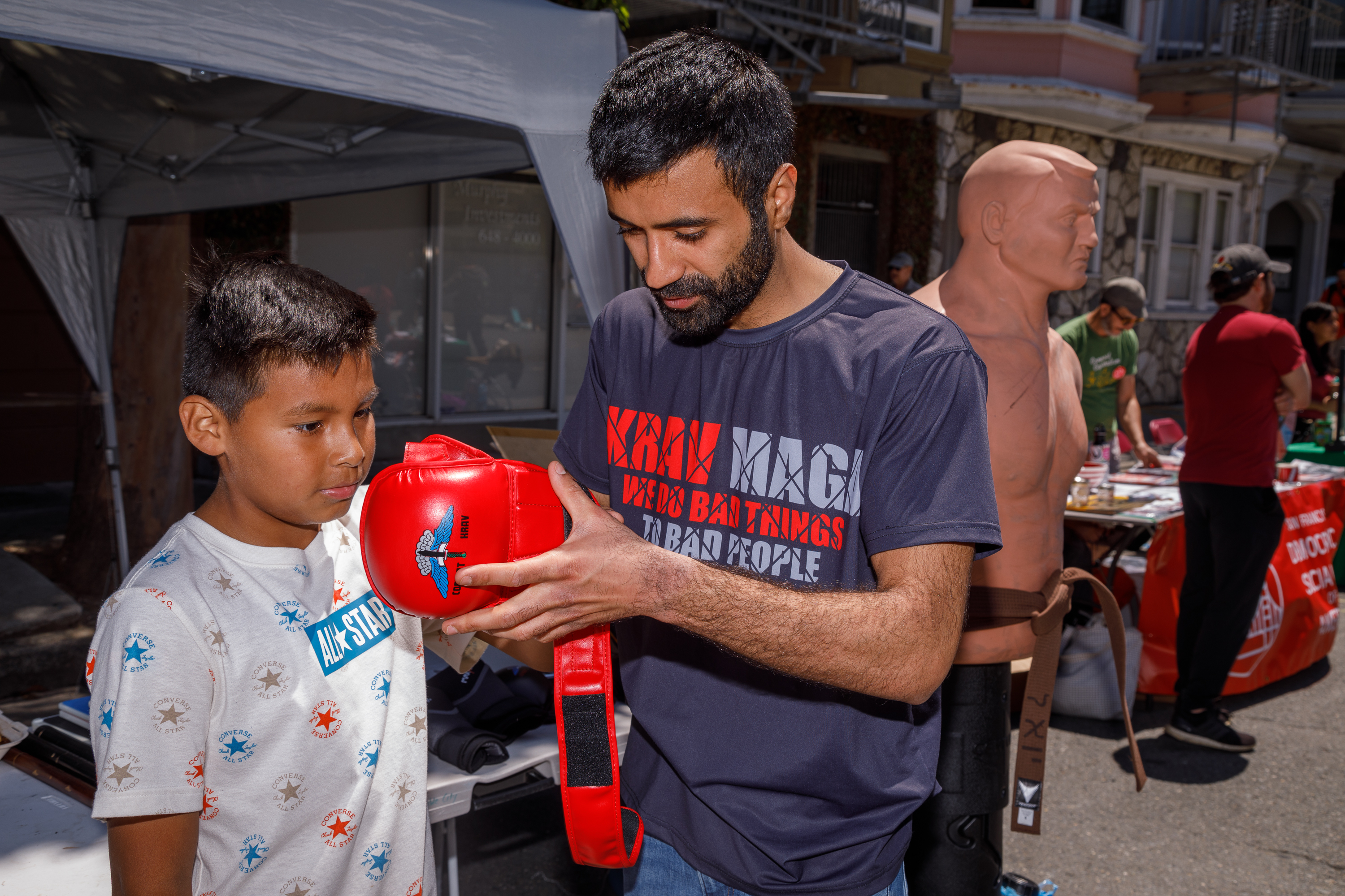 A man is showing a red boxing glove to a young boy at an outdoor stall with various items on display.