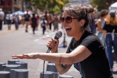 A woman with sunglasses speaks into a microphone outdoors, gesturing with her free hand. People and tents appear in the background.