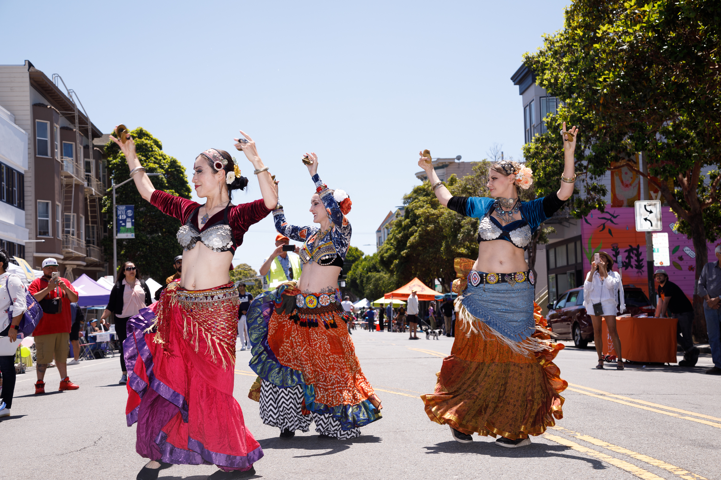 Three dancers in colorful costumes perform on a sunny street with onlookers in the background.