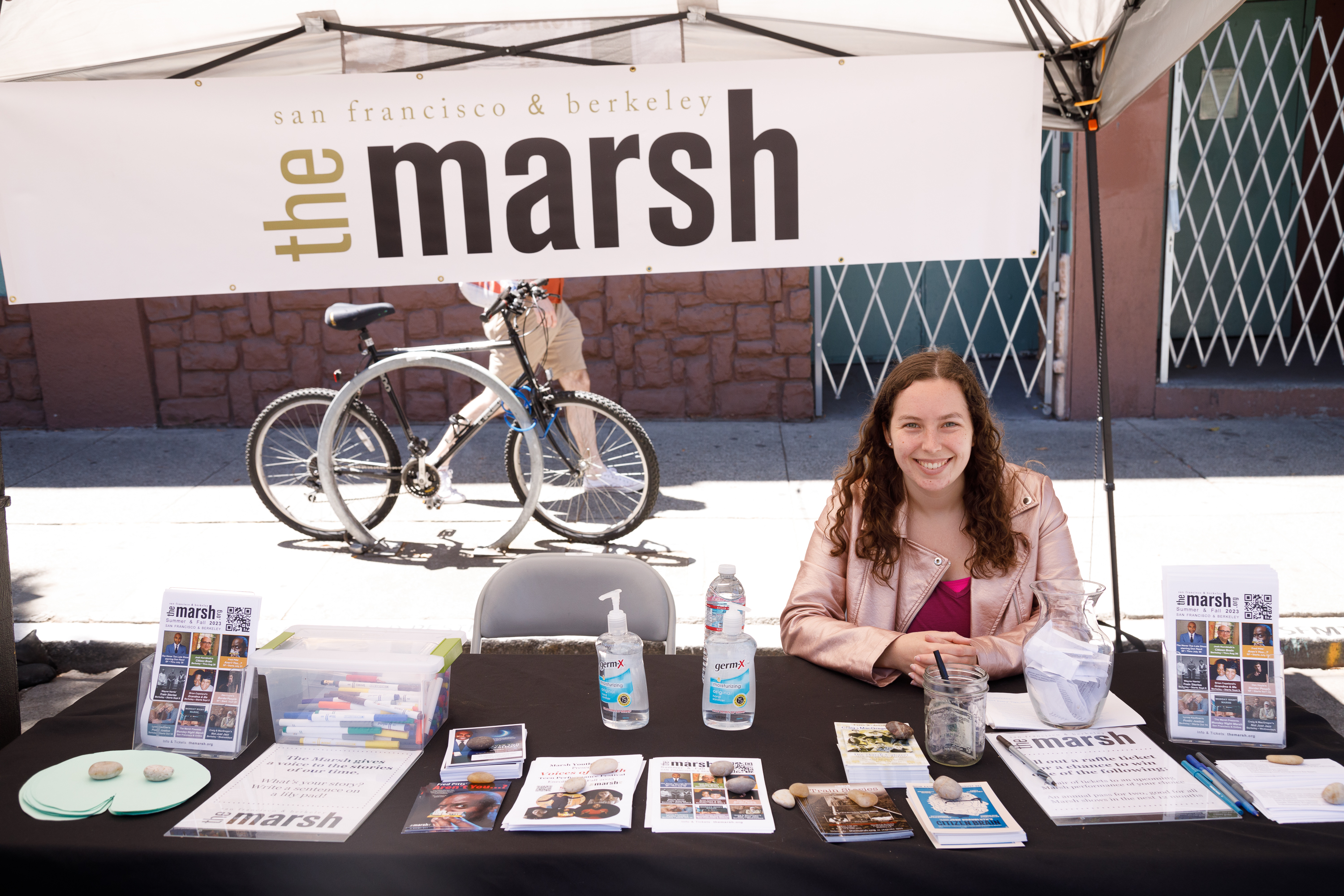 A smiling woman sits at a booth labeled &quot;the Marsh,&quot; with informational pamphlets on the table.
