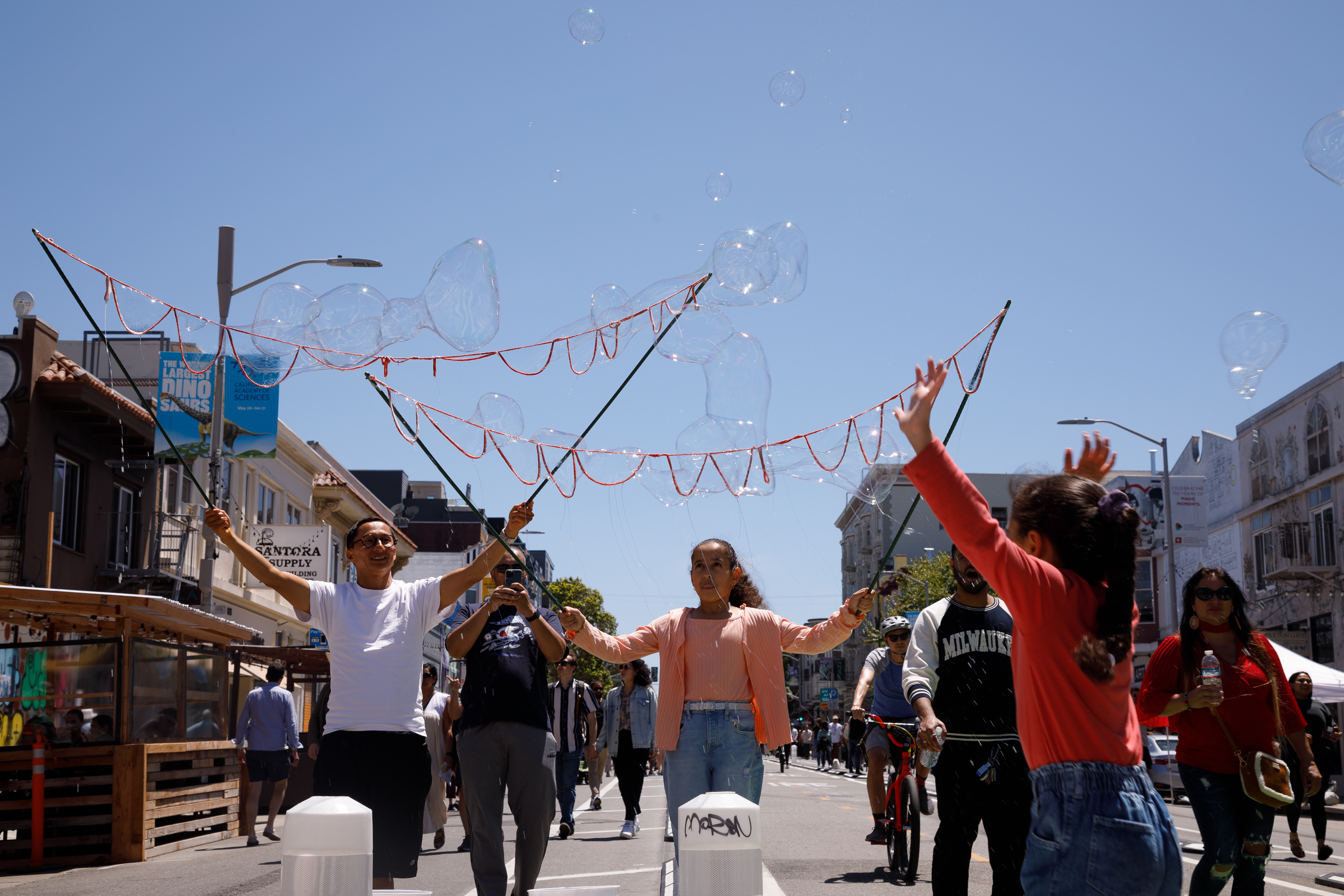People on a sunny street are creating large bubbles with a string device.