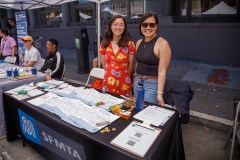 Two smiling women stand behind a booth with pamphlets, pens, and promotional items. A banner reads &quot;SFMTA.&quot;
