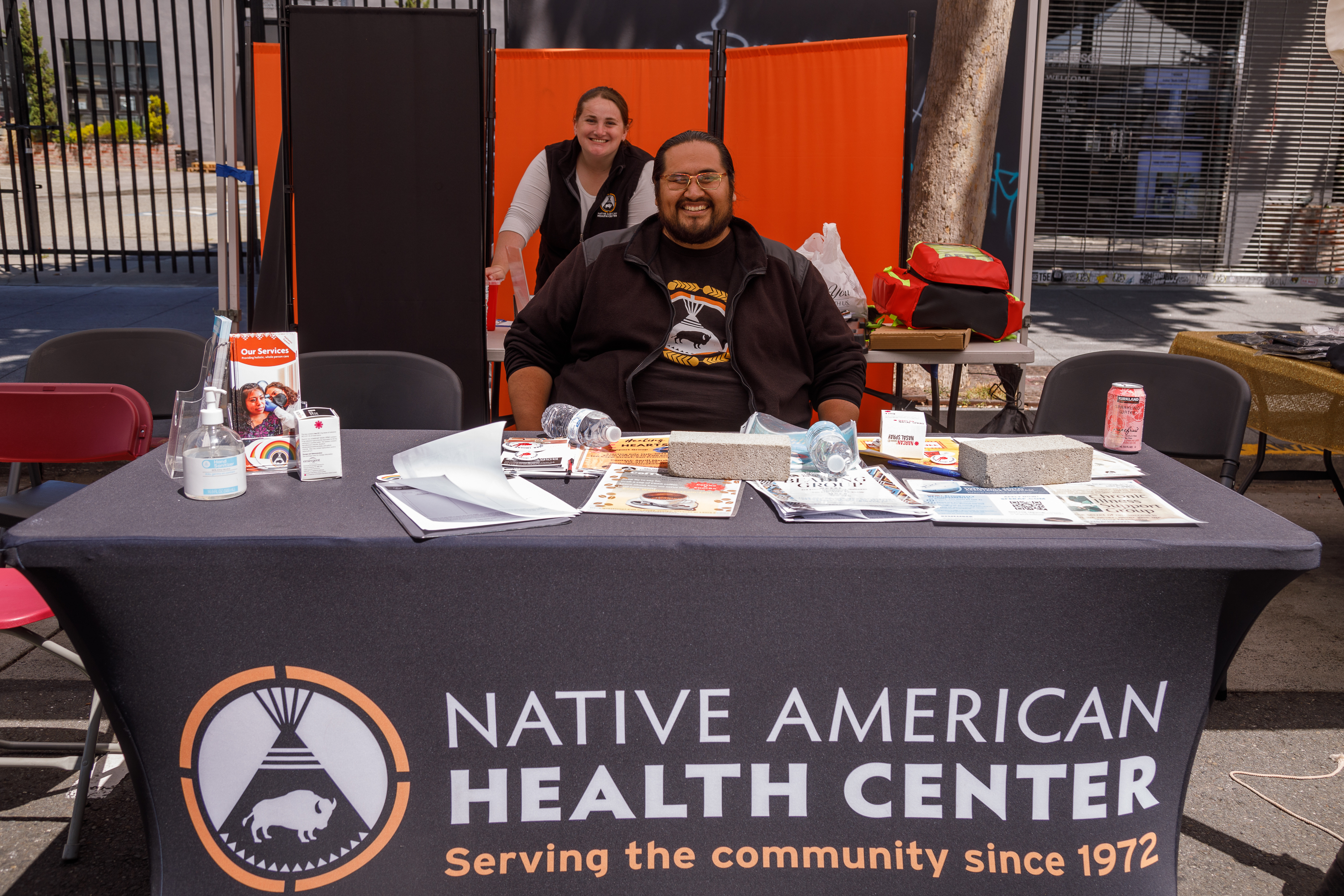 Two people staff a booth for the Native American Health Center, adorned with informational materials and a banner.