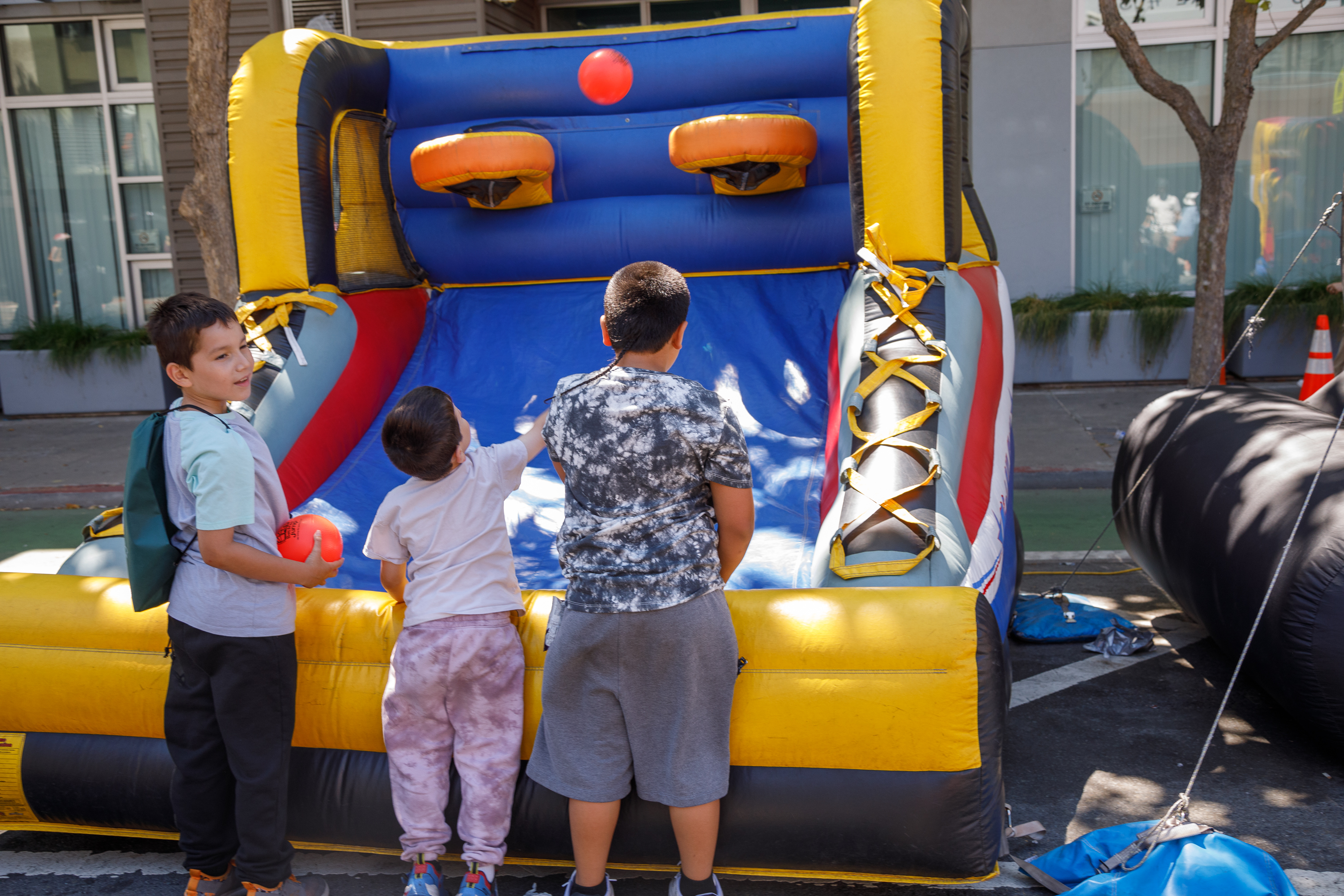 Three kids are in front of an inflatable bouncy castle, with one holding a red ball. They appear ready to play.