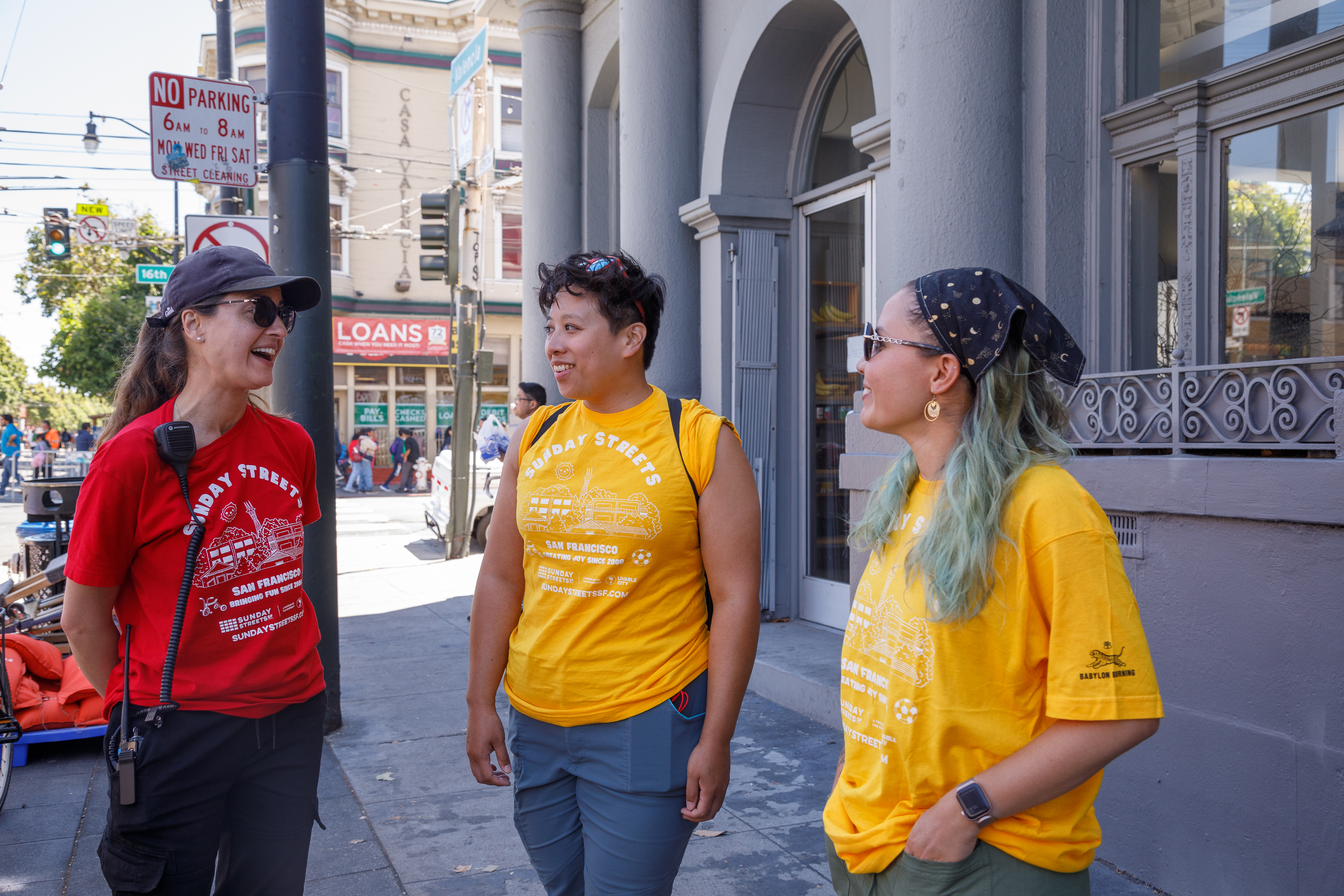 Three people in colorful t-shirts stand chatting on the sidewalk near a street corner.