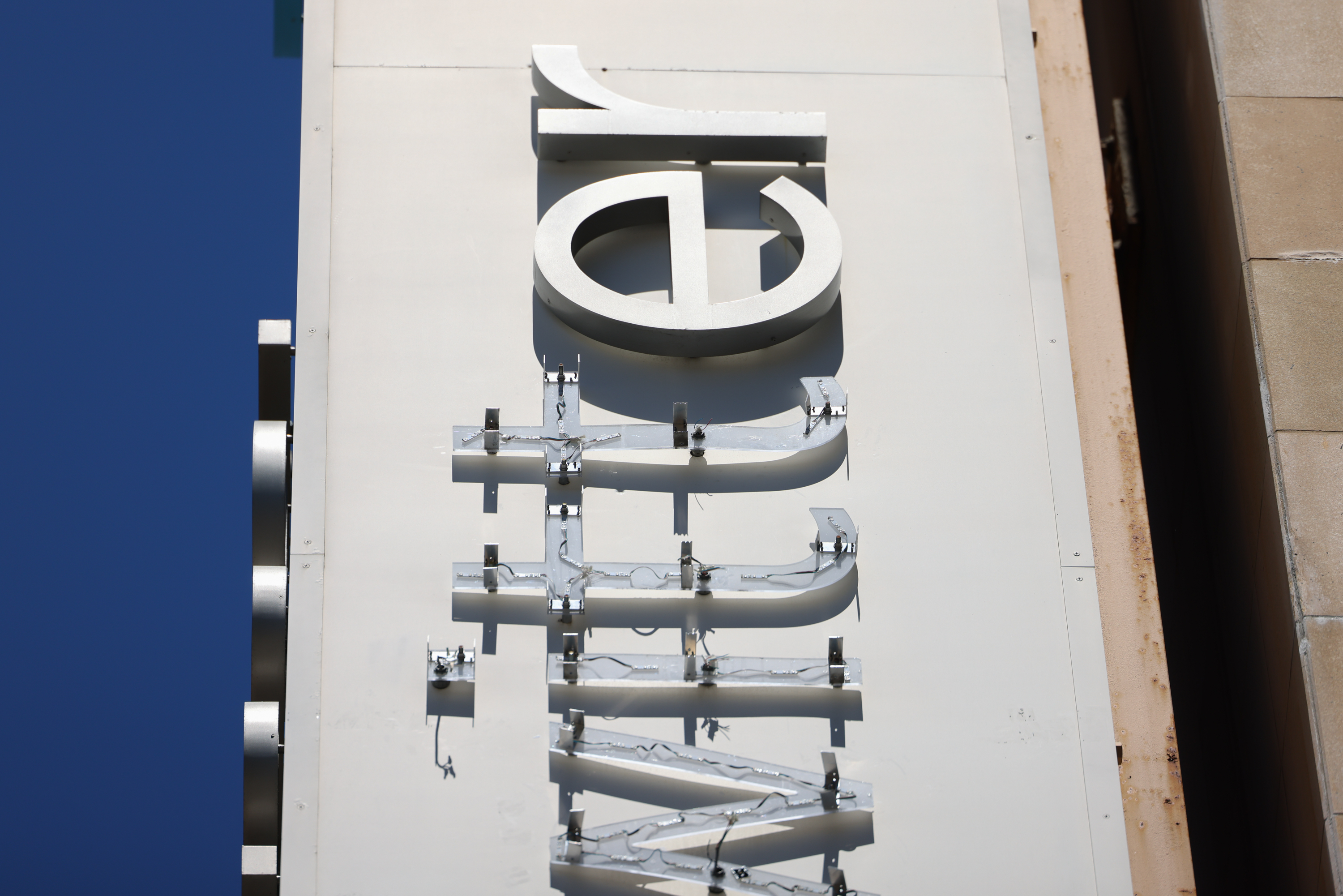 The image shows a close-up of a vertical sign on a building. The sign has detached metallic letters, possibly spelling &quot;WILLY&quot; against a clear blue sky.