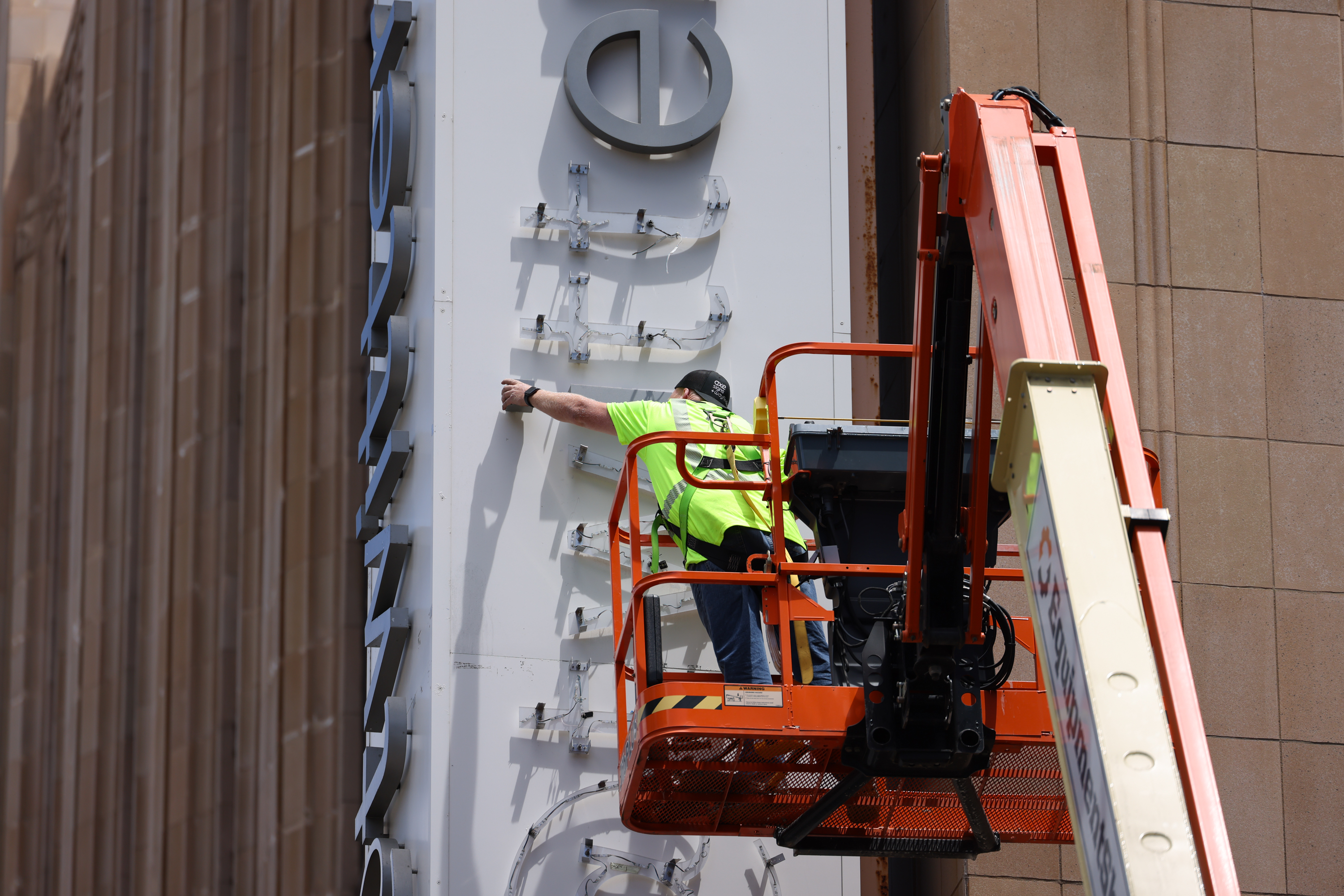 A person in a high visibility vest works on building signage from an orange aerial lift.