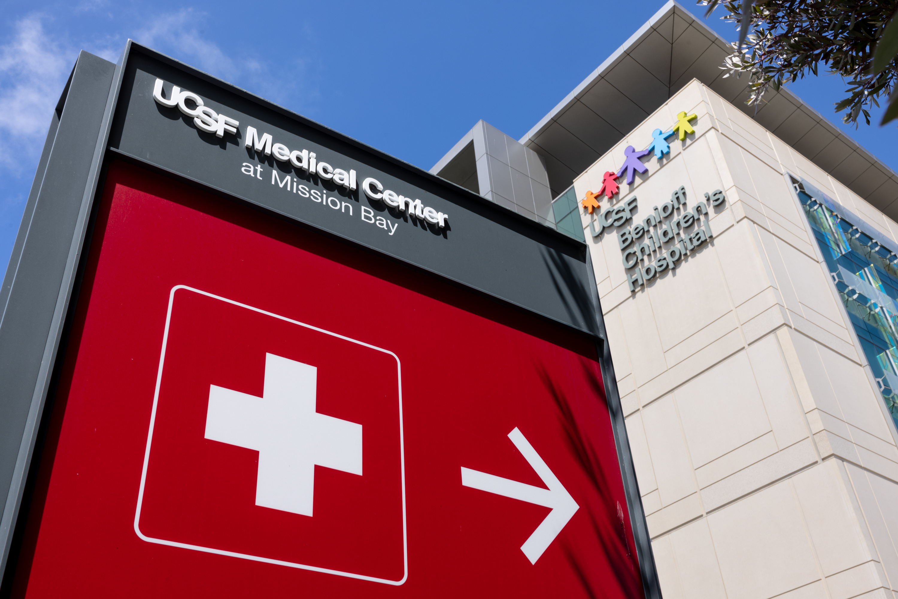 The image shows a sign for UCSF Medical Center at Mission Bay with an emergency icon and an arrow pointing right. The building includes UCSF Benioff Children's Hospital.