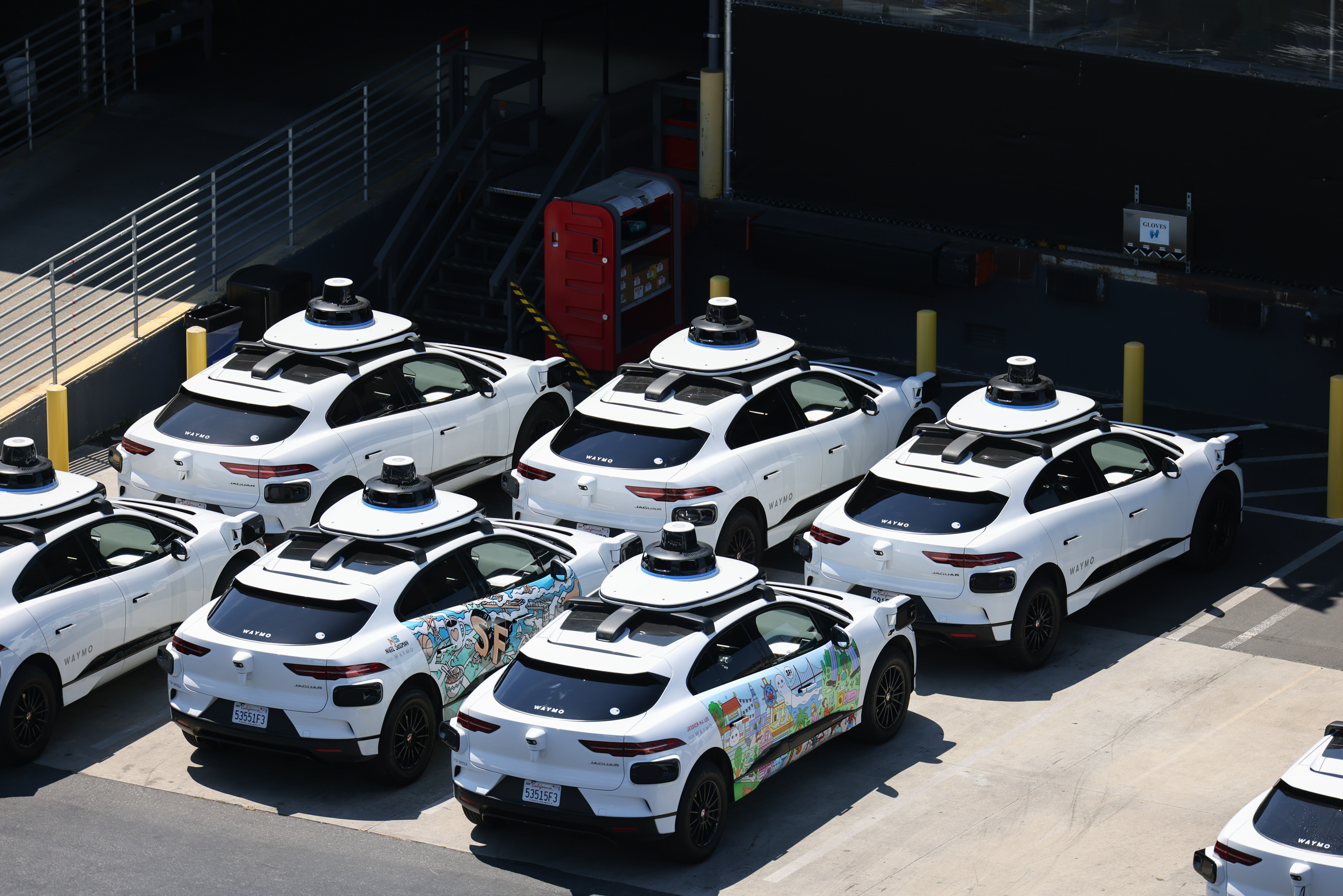 A group of white self-driving cars equipped with rooftop sensors is parked closely together in a lot, with one car featuring colorful graffiti artwork.