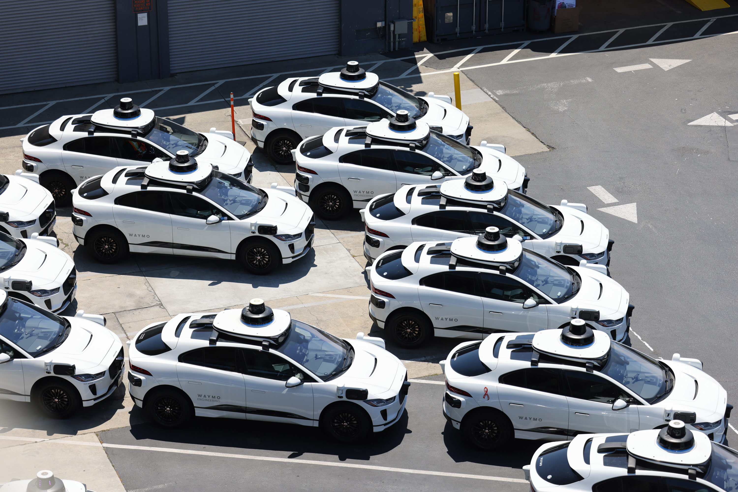 A group of white self-driving cars with rooftop sensors are parked closely together in a lot, ready for use or testing.