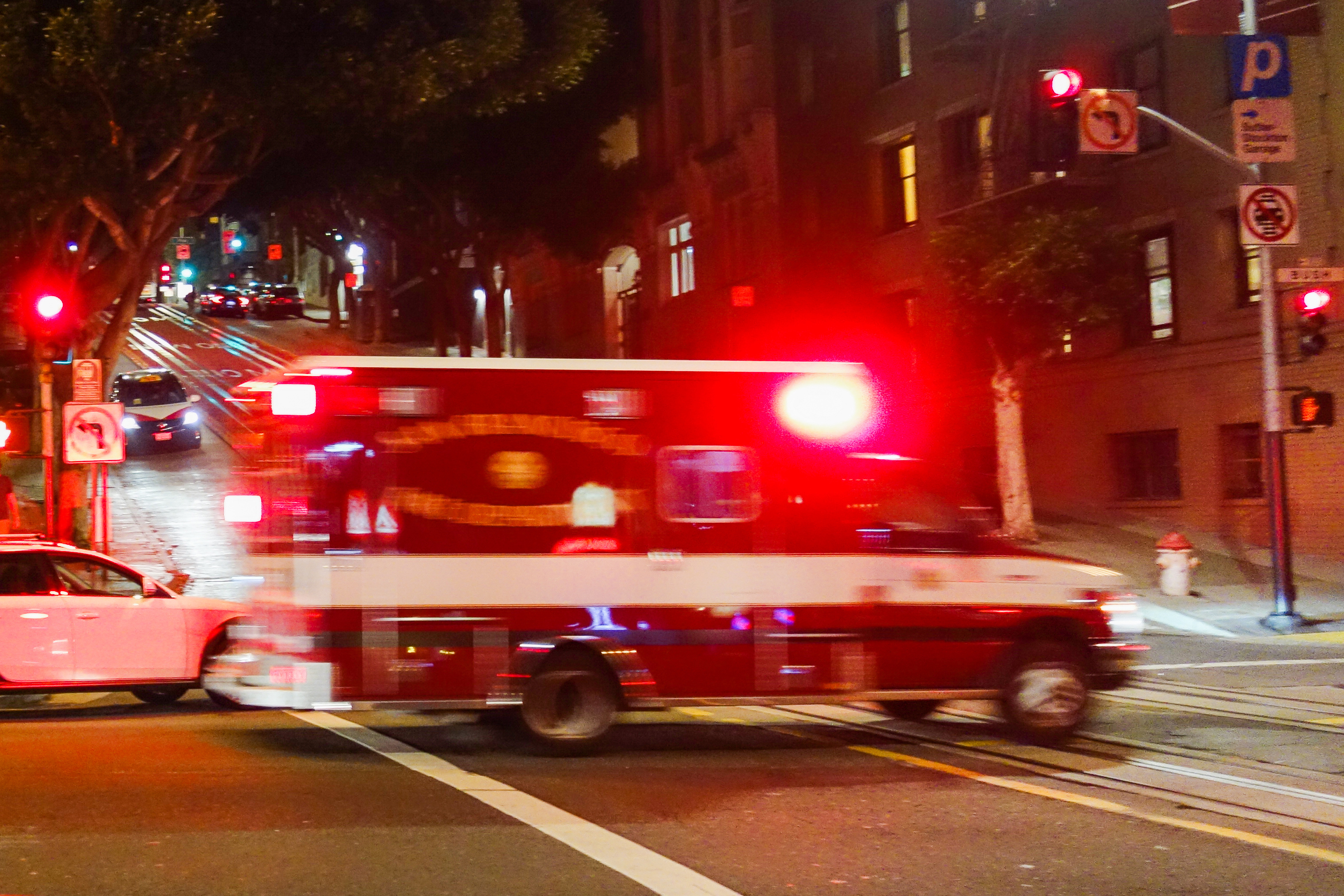 A red ambulance is seen from the side driving through an intersection with its emergency lights on.