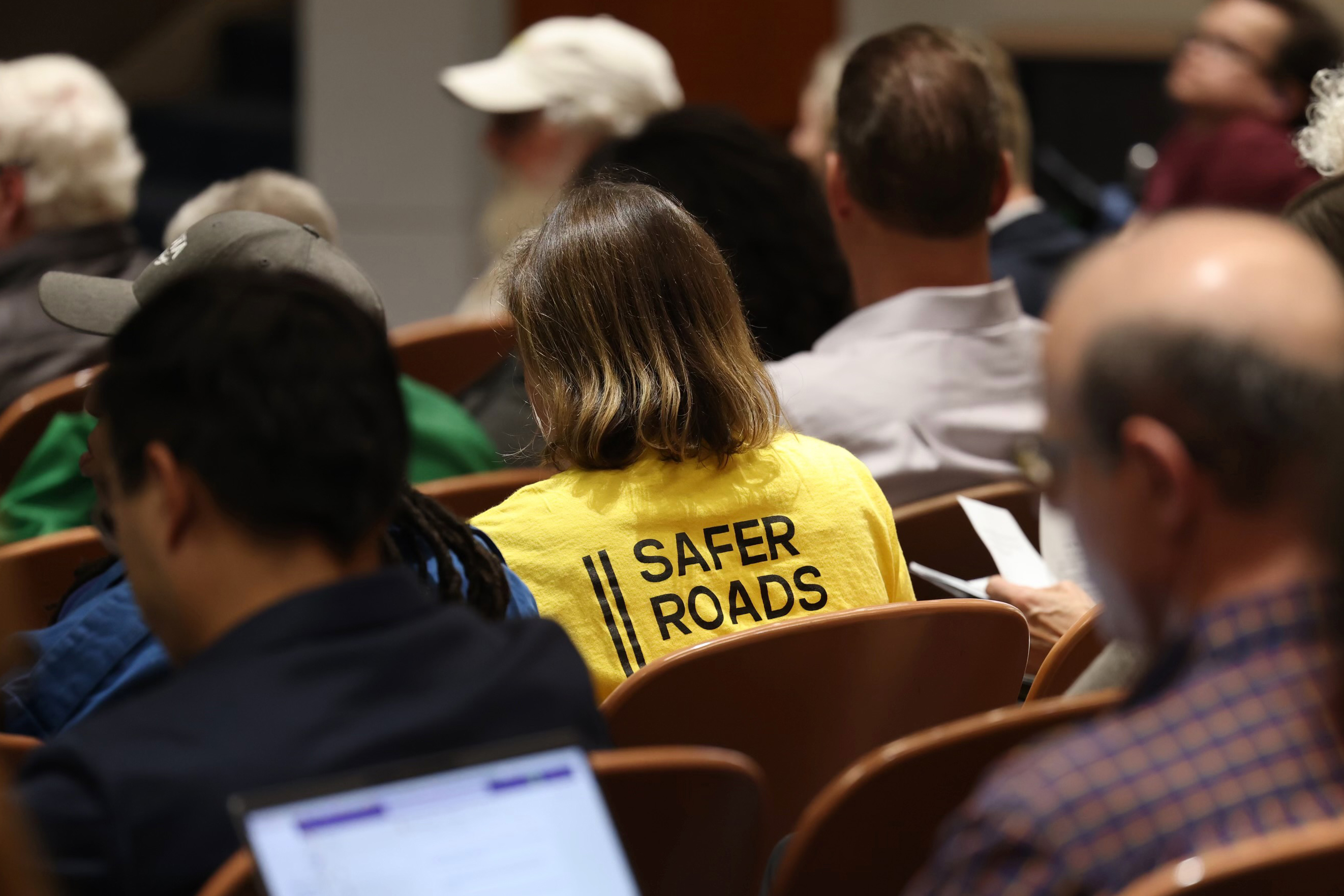 The back of an audience, with one person in a yellow shirt that reads &quot;safer roads.&quot;