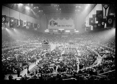 A crowded indoor arena with banners, stage in center, spectators, and a &quot;FOR THE PEOPLE&quot; slogan.