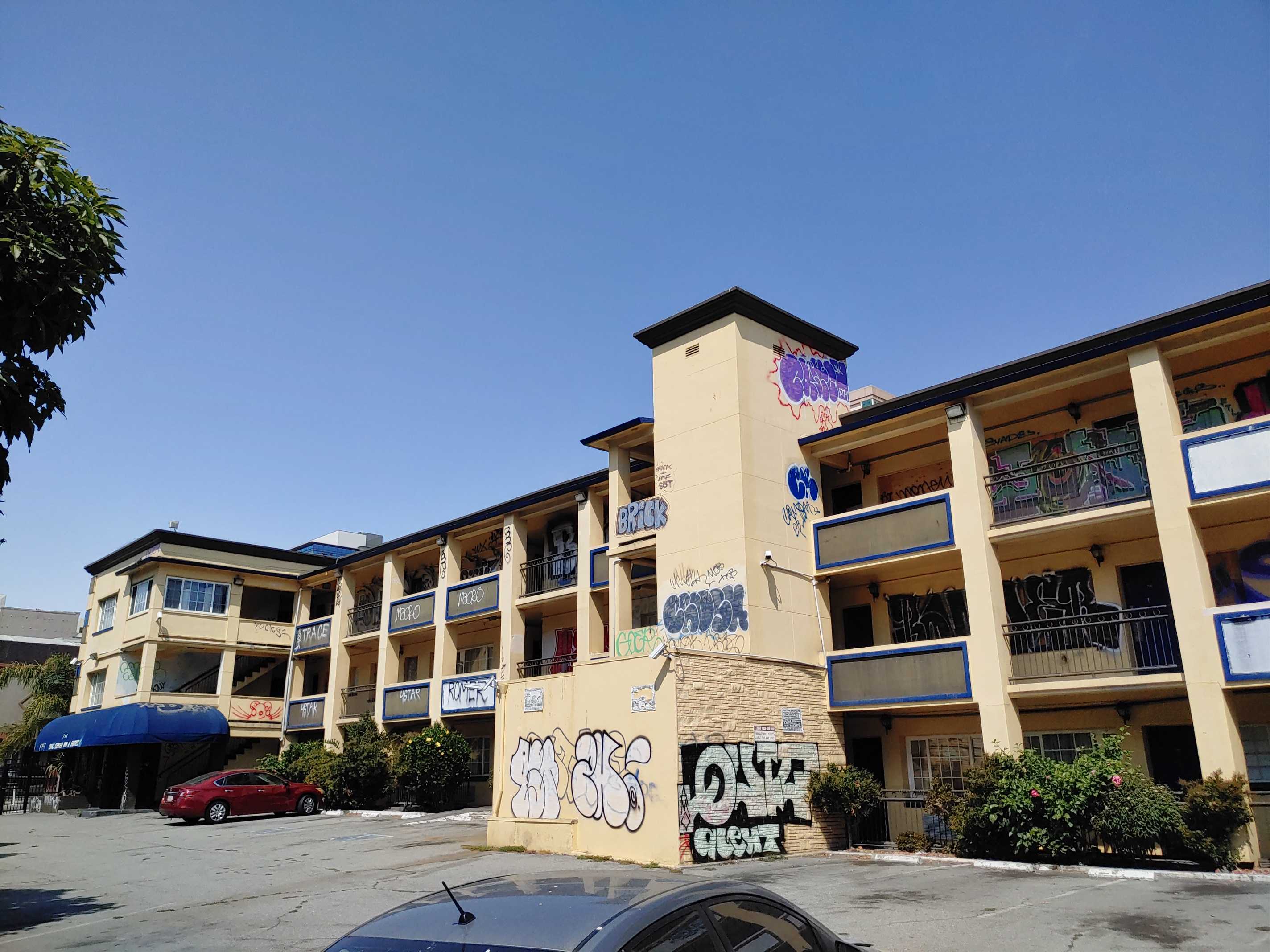 A three-story motel with graffiti on its walls under a clear blue sky. A red car is parked in the lot.
