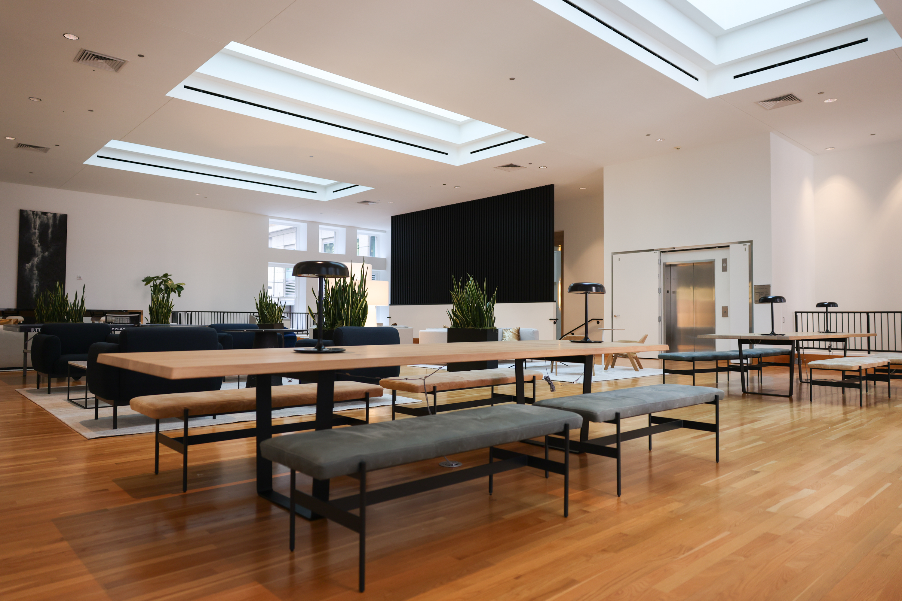 The image shows a spacious, modern room with wooden floors, long tables, benches, plants, and skylights. The design is minimalistic with ample seating.