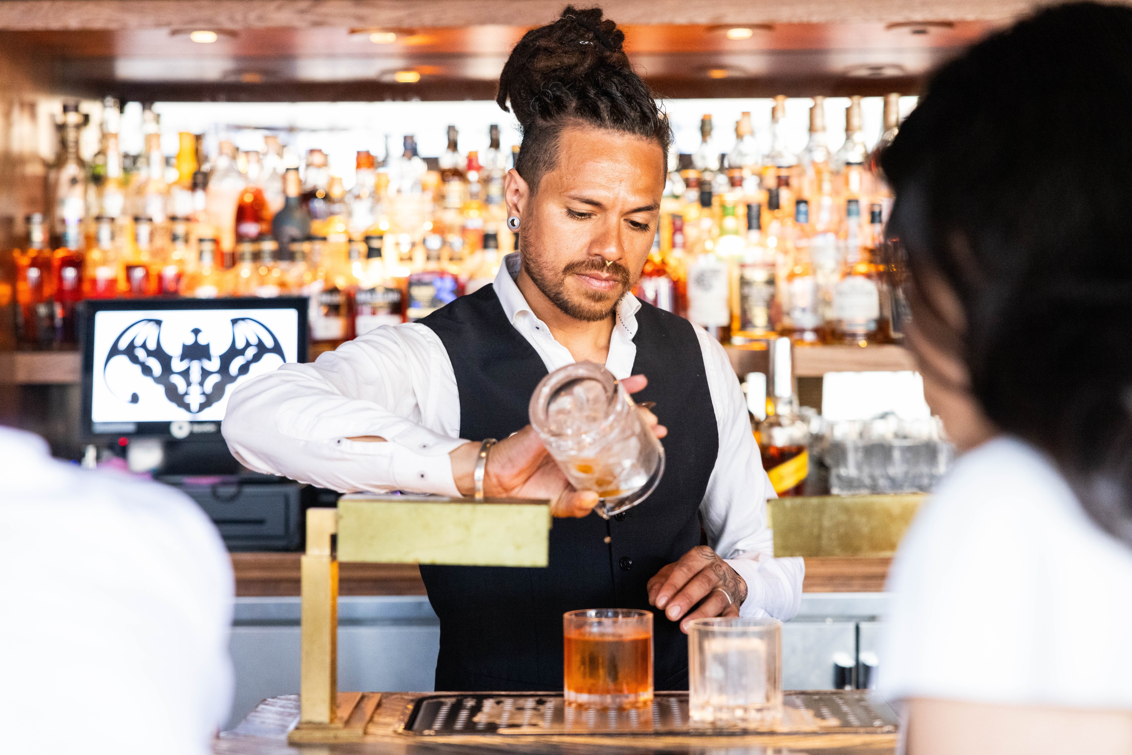 A bartenders strains two drinks on a golden bar with a row of bottles behind him. 