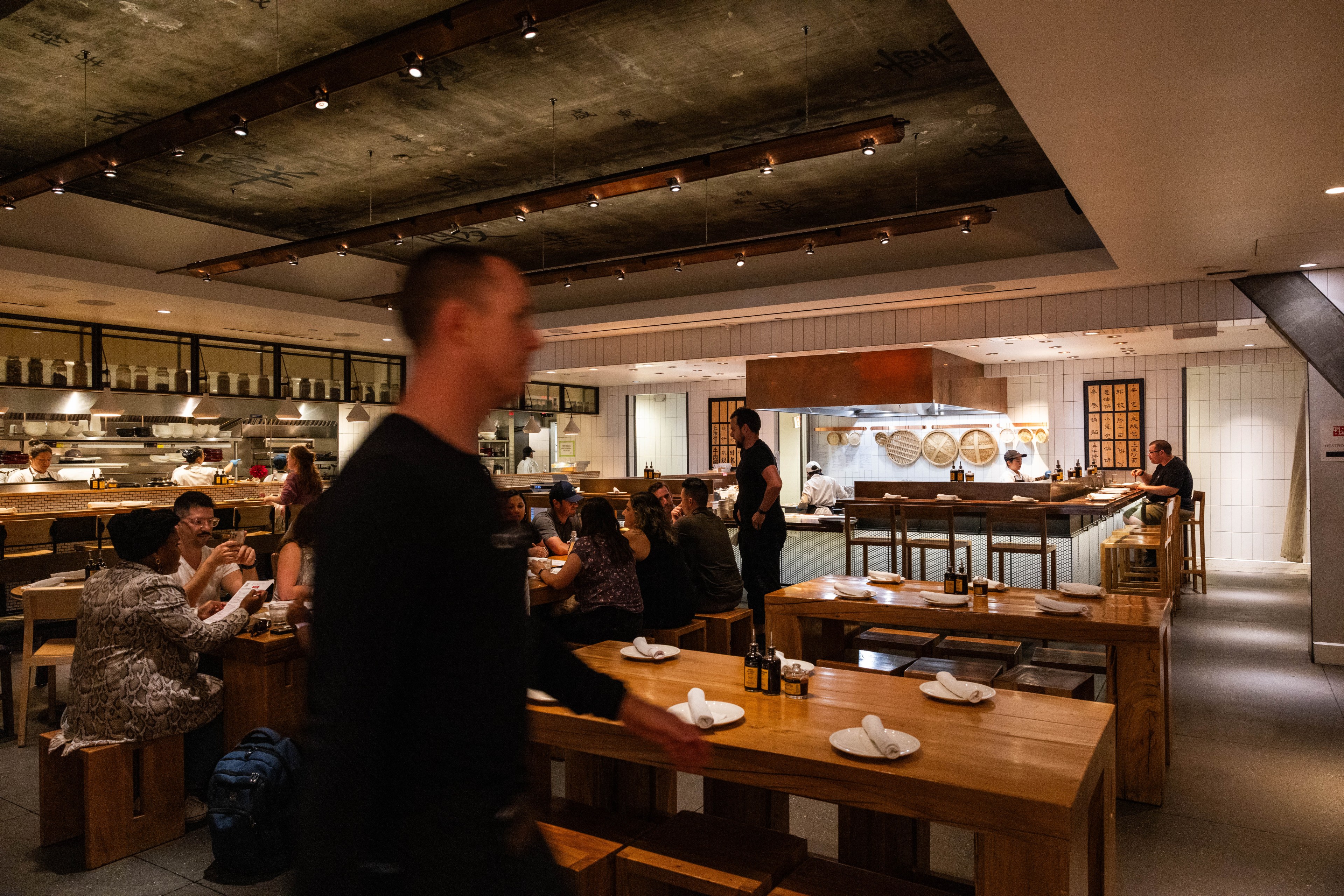 A bustling restaurant interior with patrons dining, an open kitchen, and a blurred figure walking by.