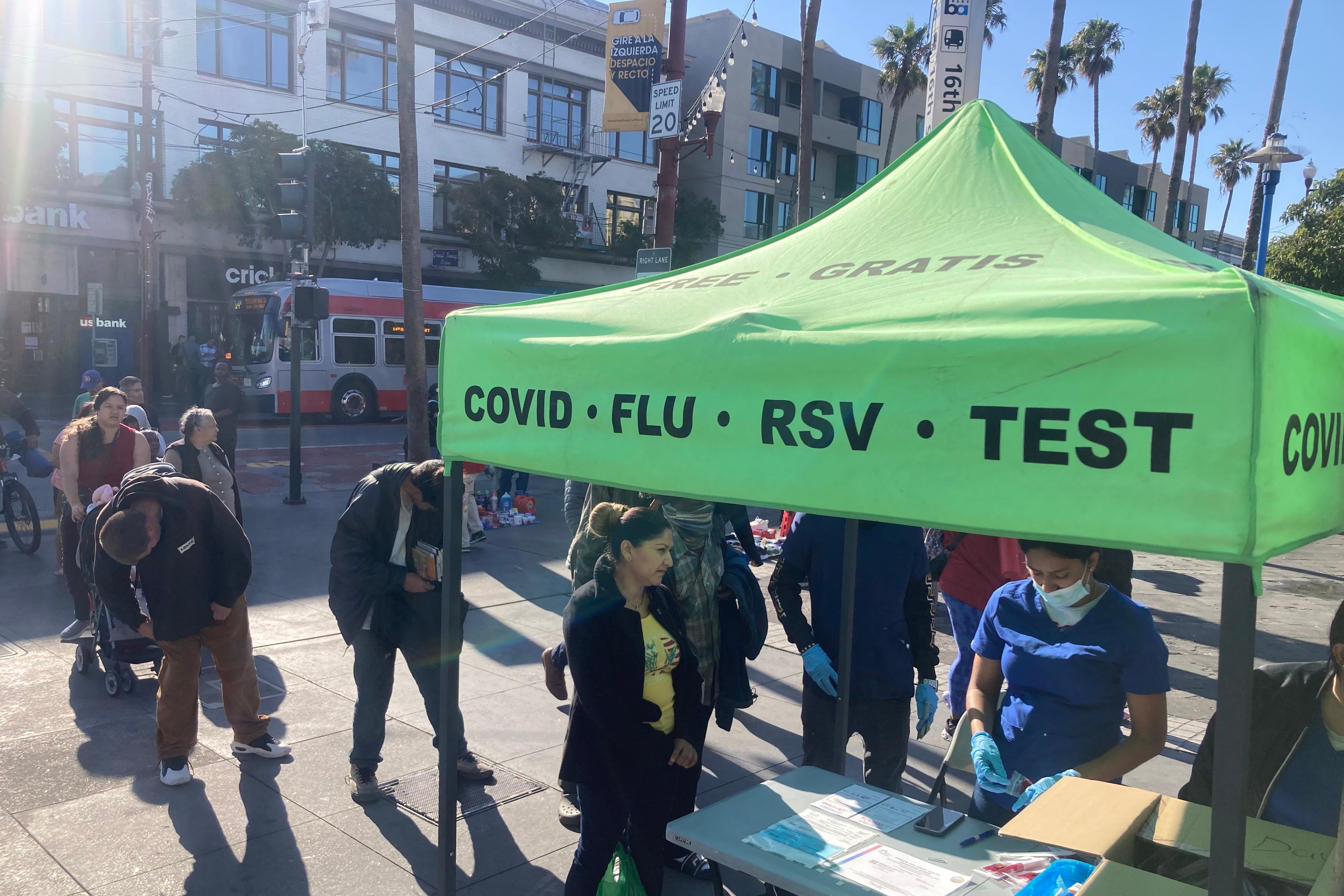 A green pop-up tent with the words COVID, FLU, RSV and TEST on its edge shelters people on a sunny day.