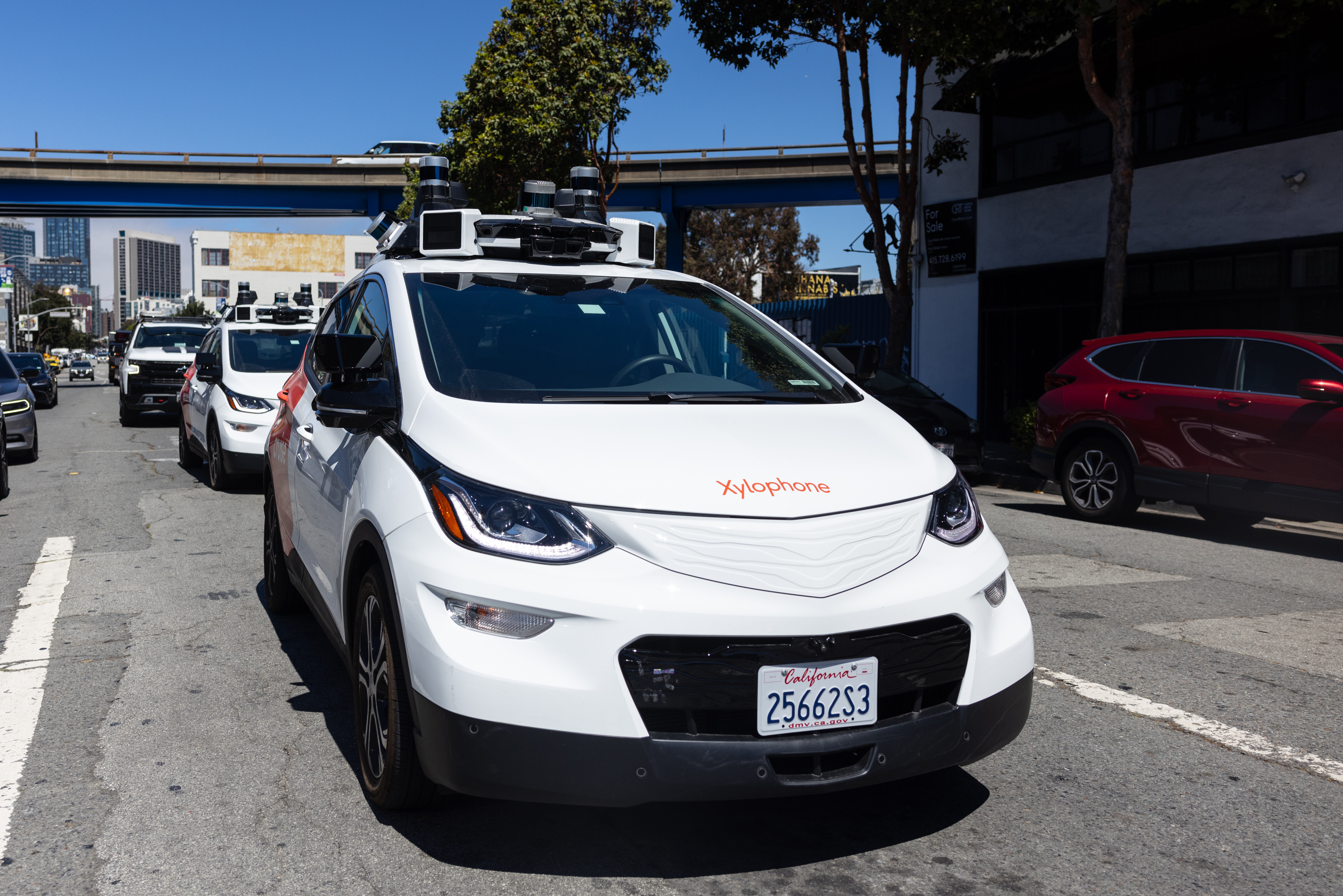 A white autonomous car with sensors on the roof drives on a city street. The car has "Xylophone" on the front, with a building and trees in the background.