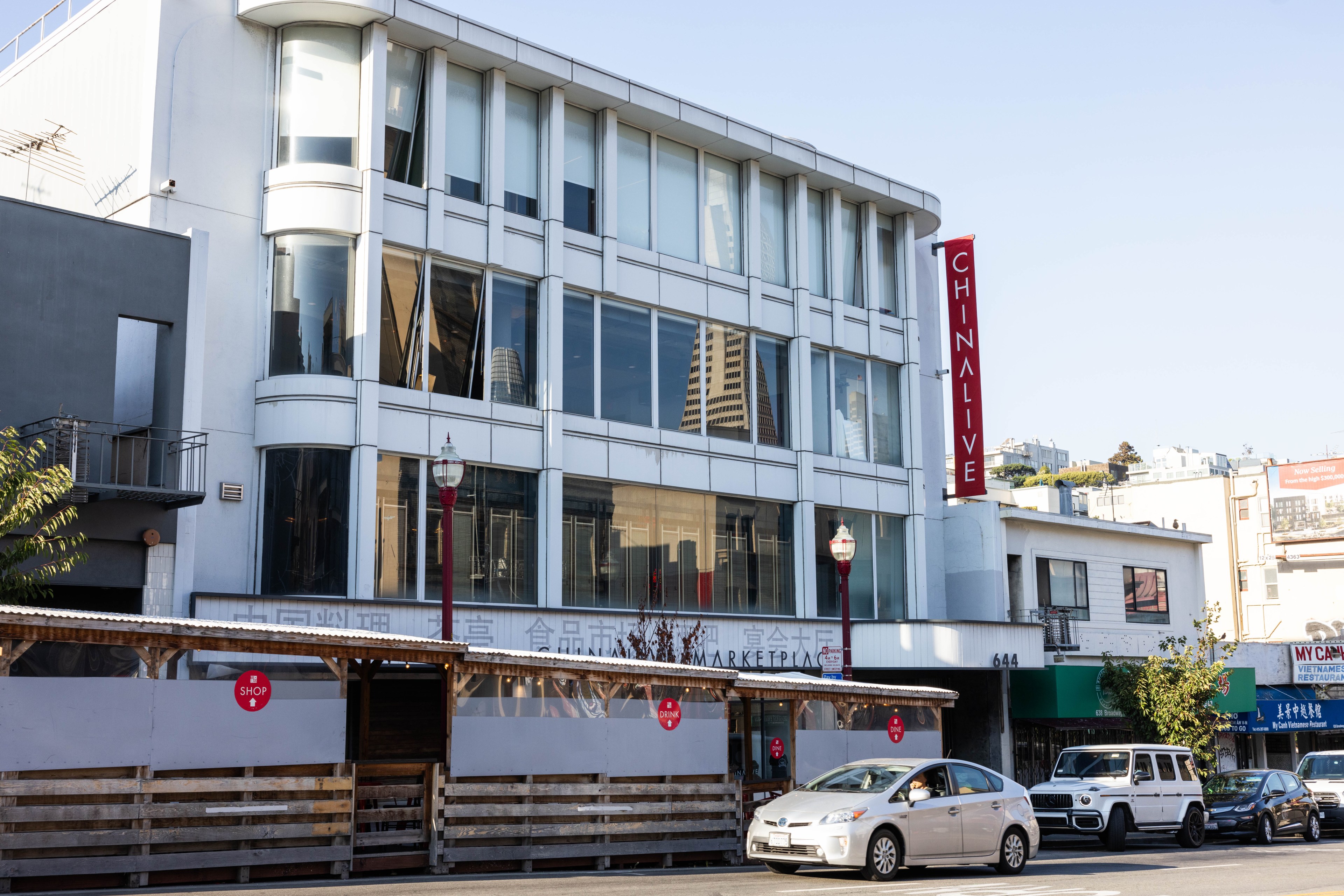 A modern three-story building with large windows displays a vertical &quot;China Live&quot; sign. It's beside a street with cars and nearby smaller shops.