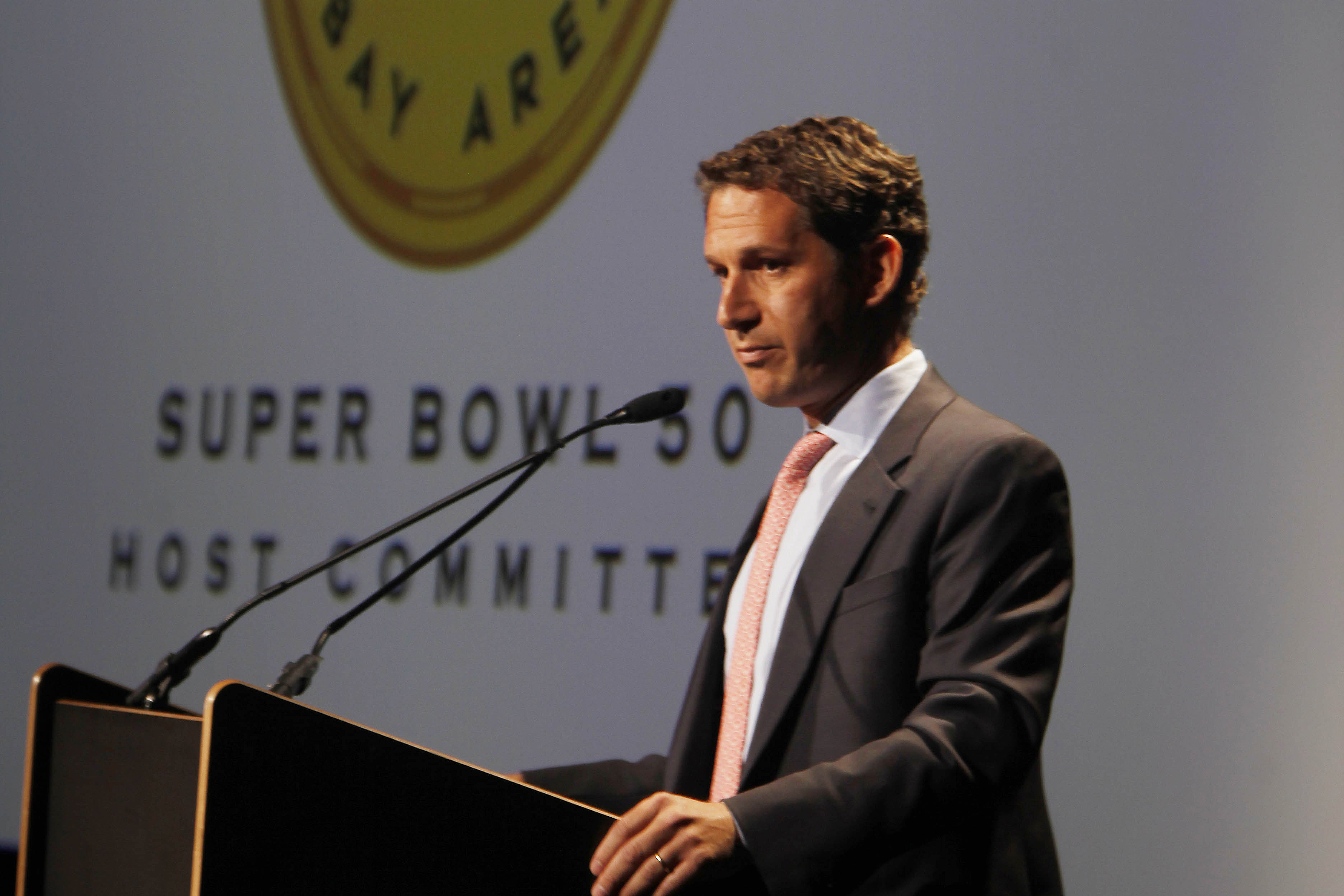 A man in a suit speaks at a podium with the "Super Bowl 50 Host Committee" logo in the background.