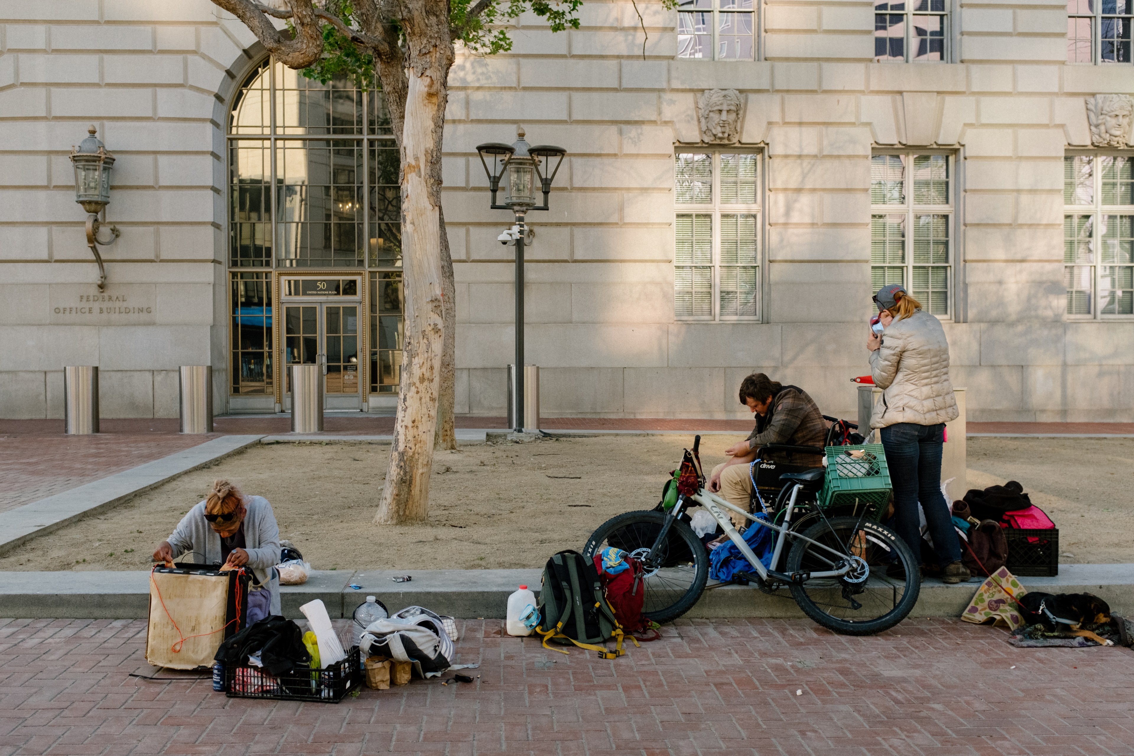 Three people sit by a building with belongings, a bicycle, and a dog lying down.