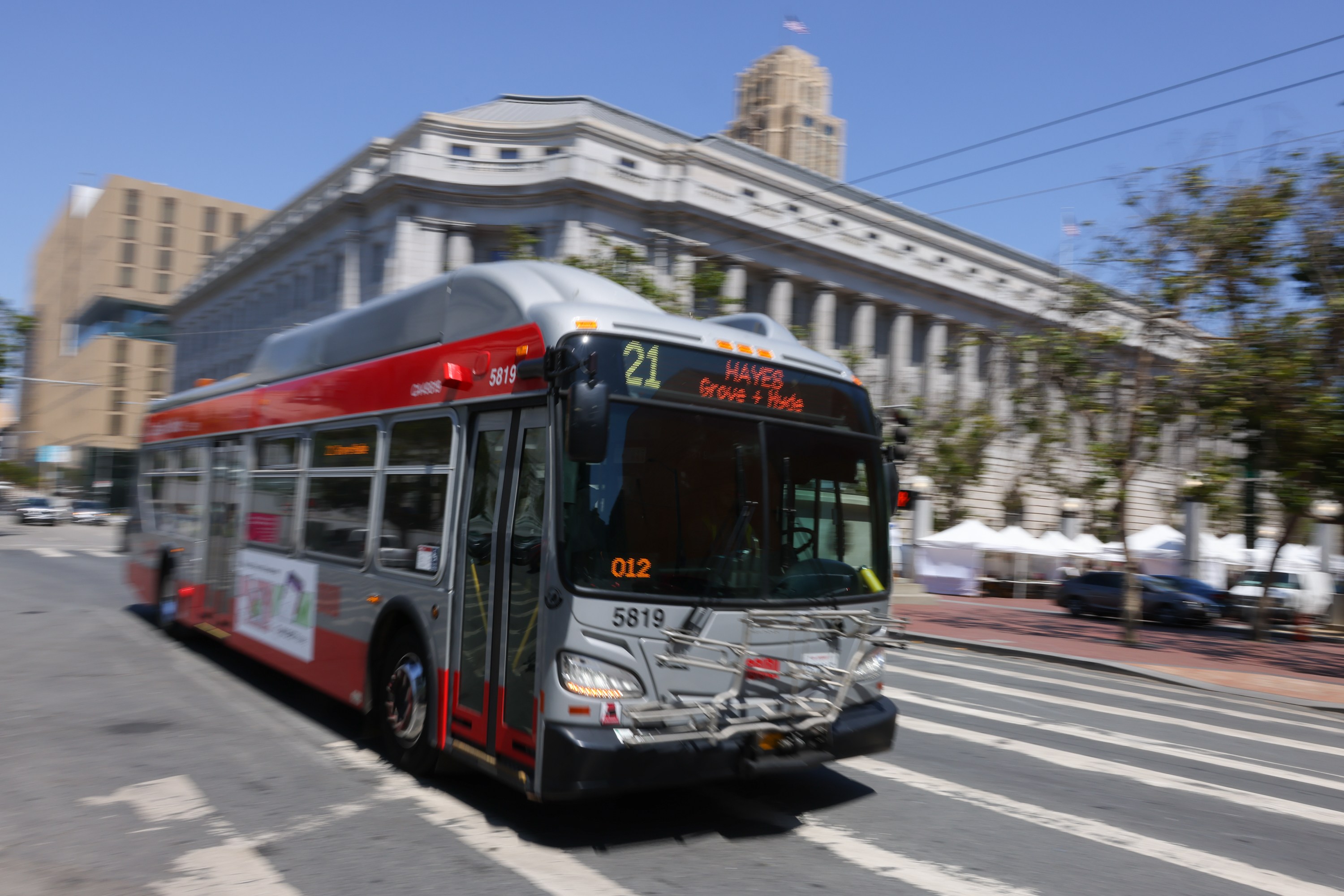 A gray and red city bus, marked route 21 to Hayes, moves along a crosswalk. It passes a large, classical-style building on a sunny day.