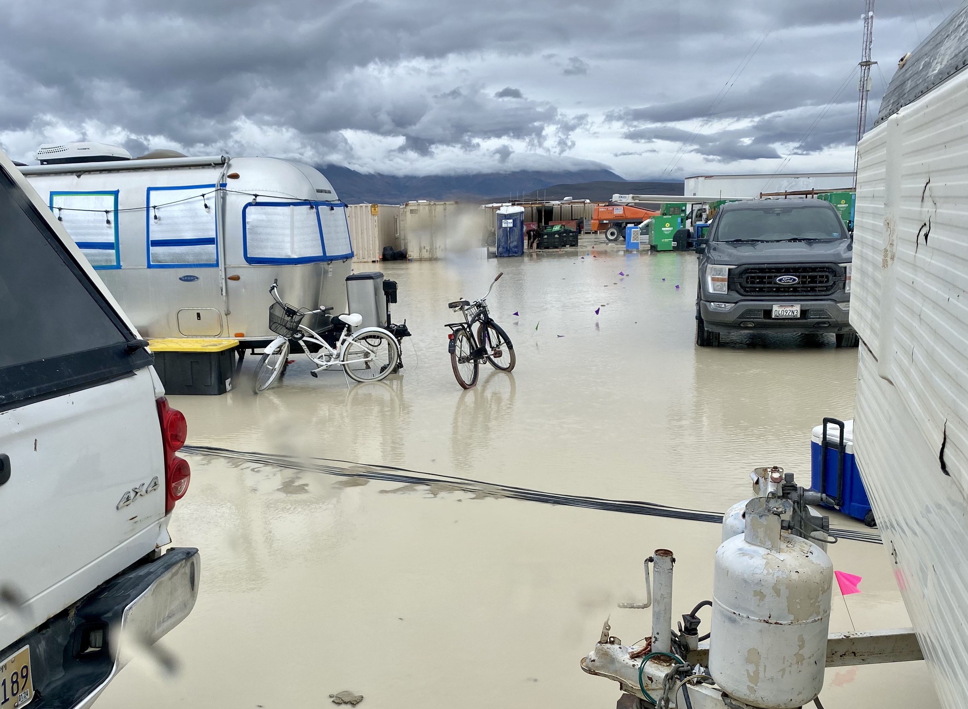 A flooded campground with vehicles, trailers, and submerged bikes, under stormy skies.