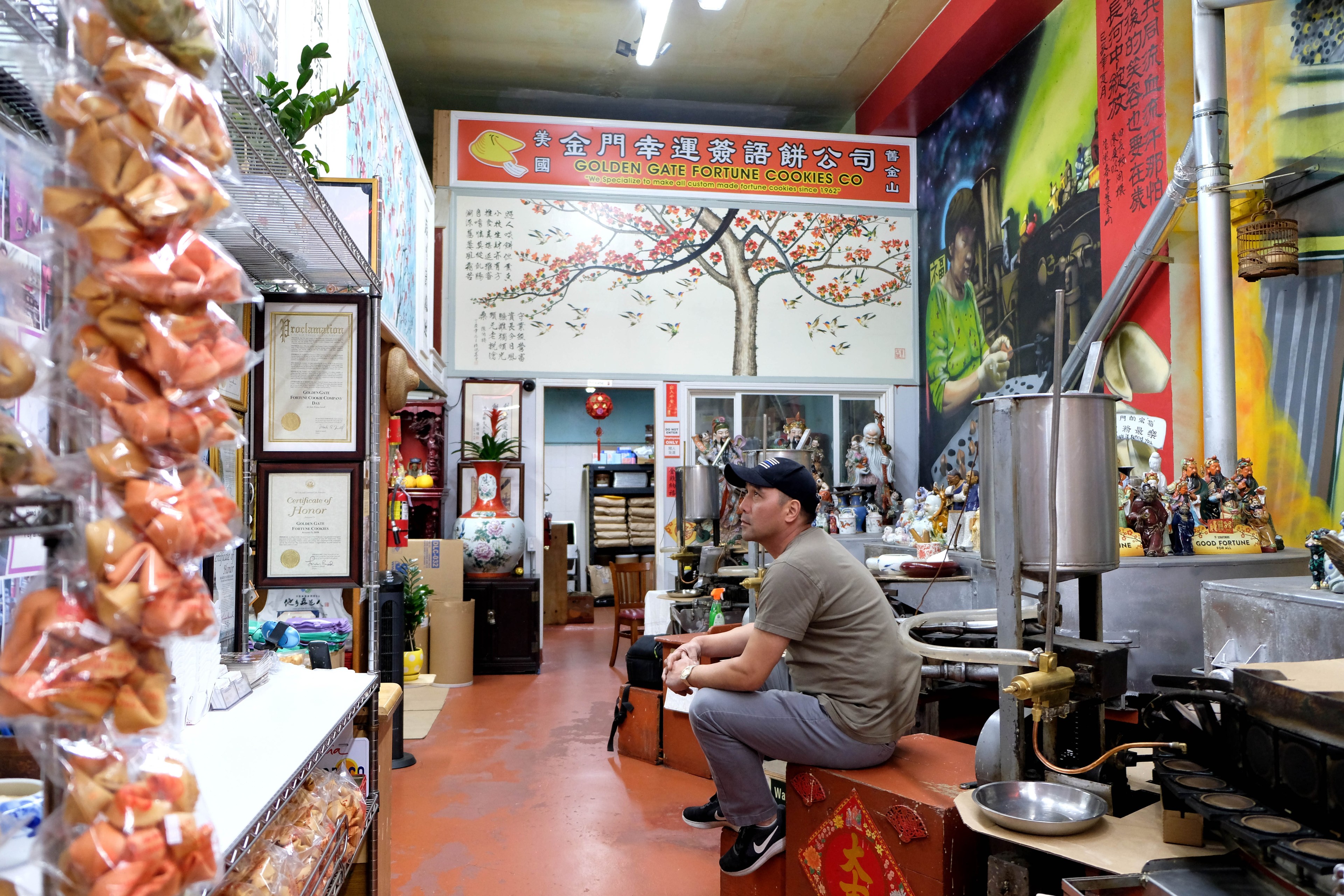A man sits in a tiny factory space that has fortune cookie making equipment and bags of fortune cookies.