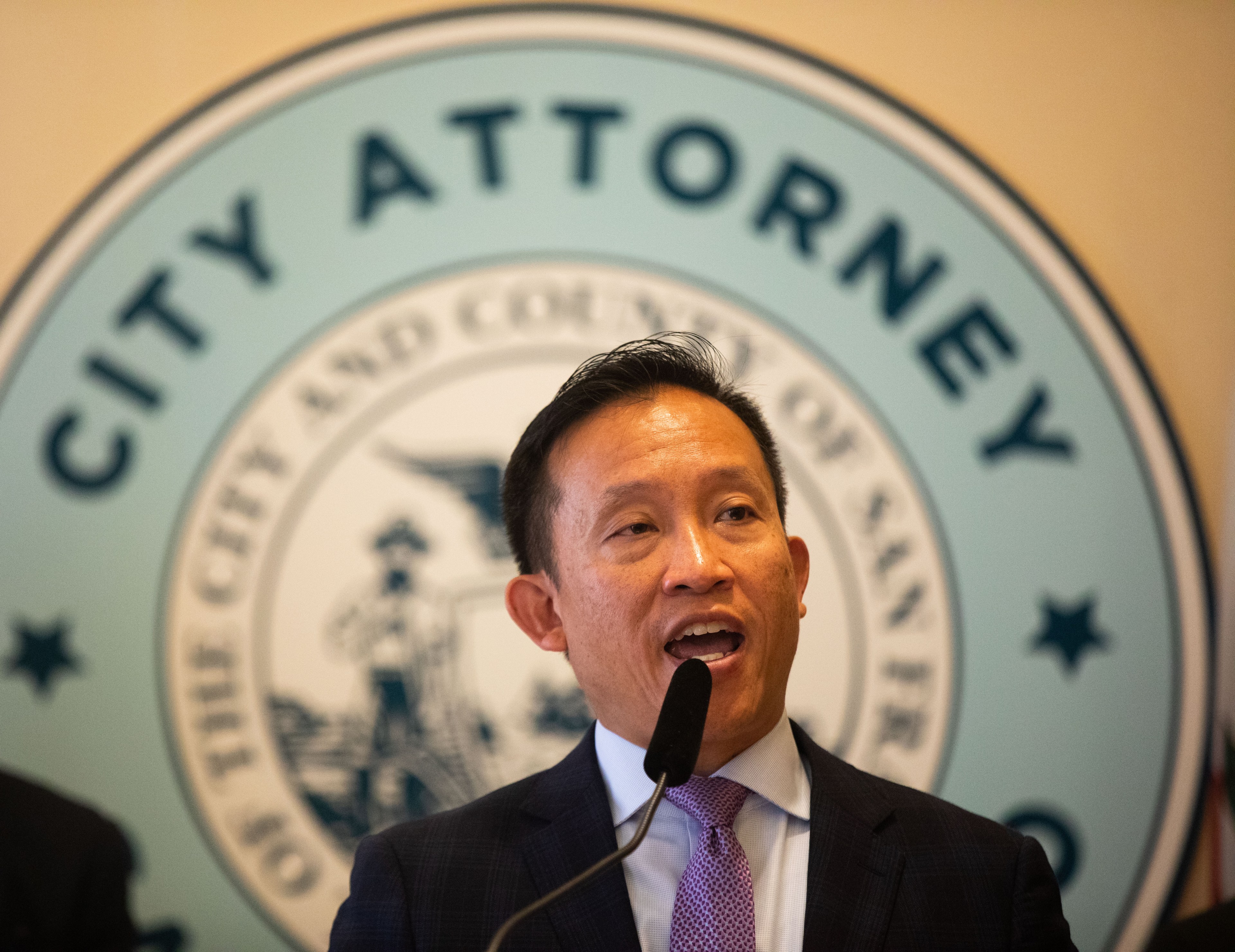 A man in a suit and tie speaks at a microphone, in front of a sign that reads "City Attorney" and features the seal of San Francisco.